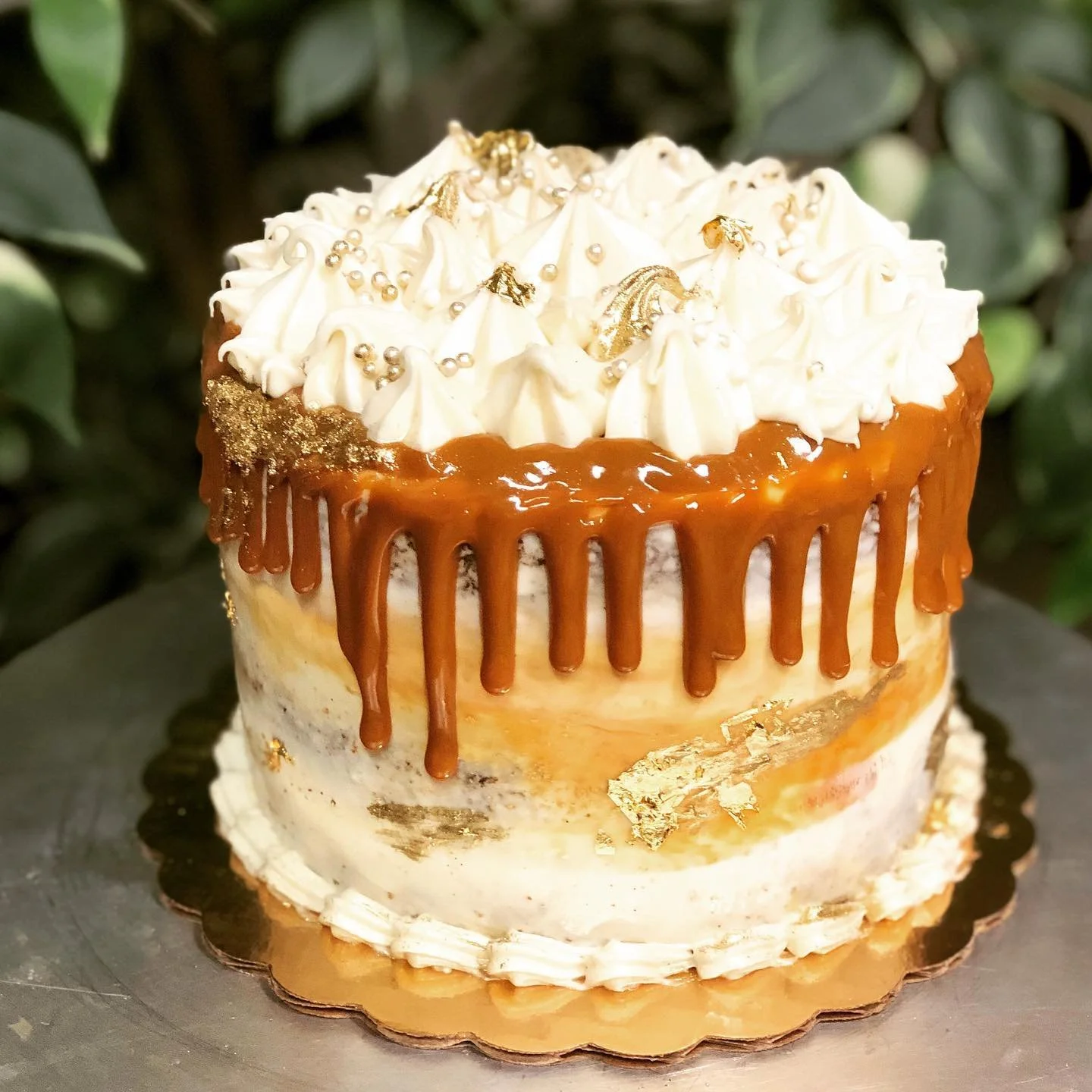 Decorated birthday cake with white frosting, gold and silver sprinkles, gold leaf, and caramel drip on top, sitting on a golden cake board.