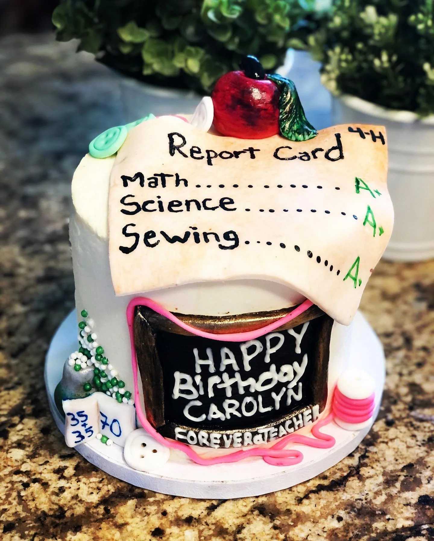 A birthday cake shaped like a school report card with messages and decorations. The report card has subjects like math, science, and sewing, with grades A and A. The cake features a red apple on top, green leaves, a chalkboard with the message "Happy