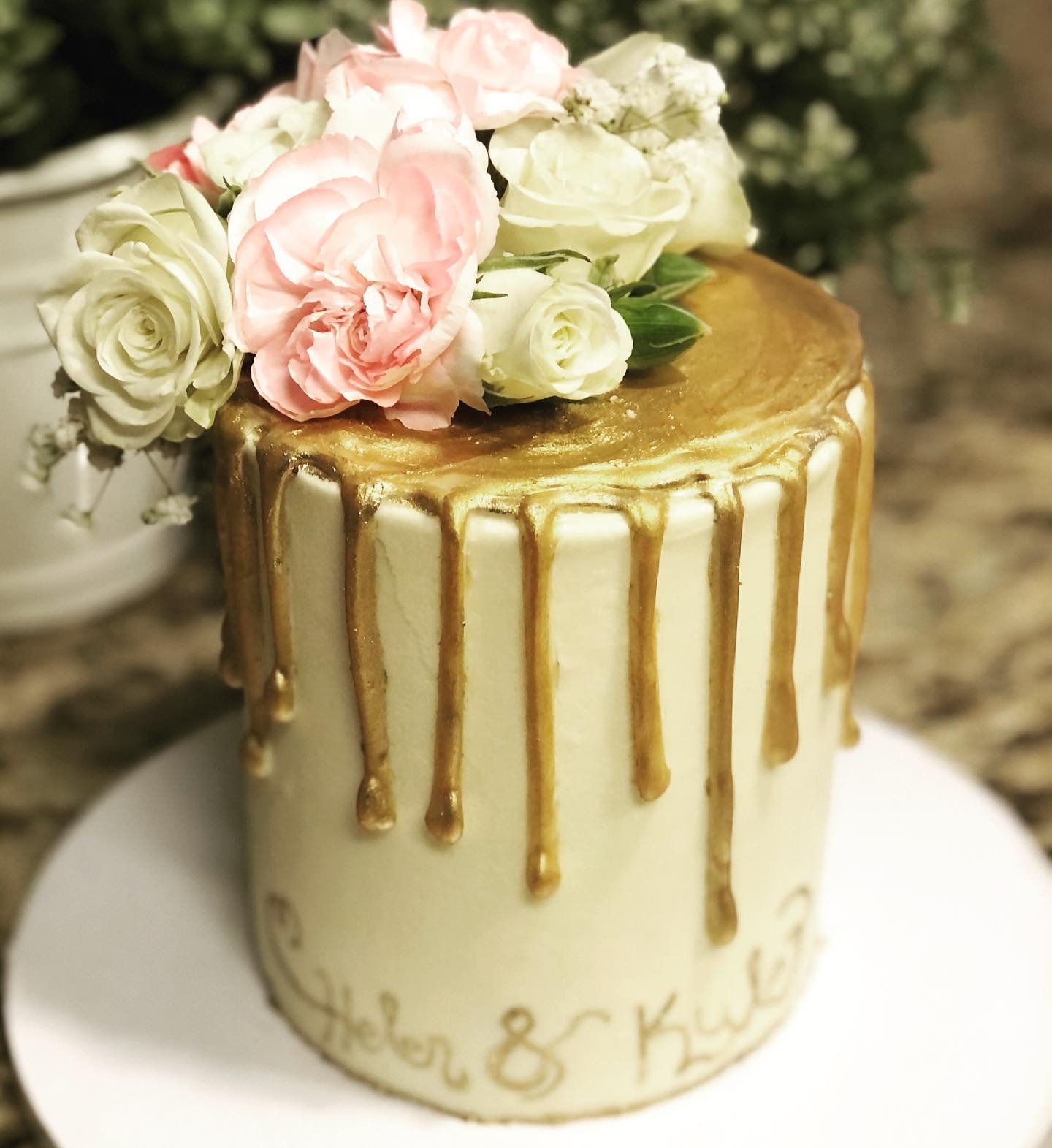 A decorated cake with gold drip icing and pink and white flowers on top, sitting on a white plate.