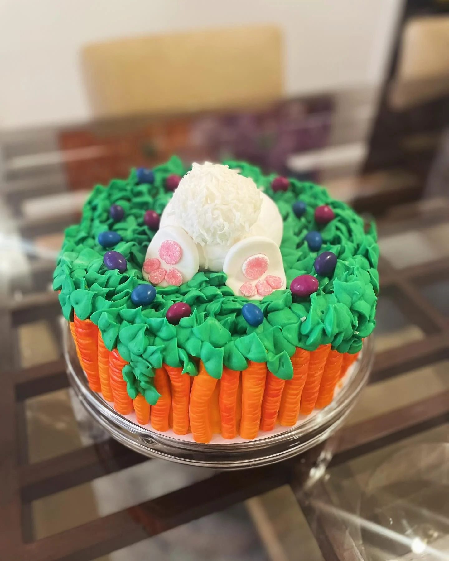 A cake decorated with orange carrots around the base, green icing resembling grass on top, and a white edible decoration in the shape of a train with a white spherical element on top, topped with small colorful candies.