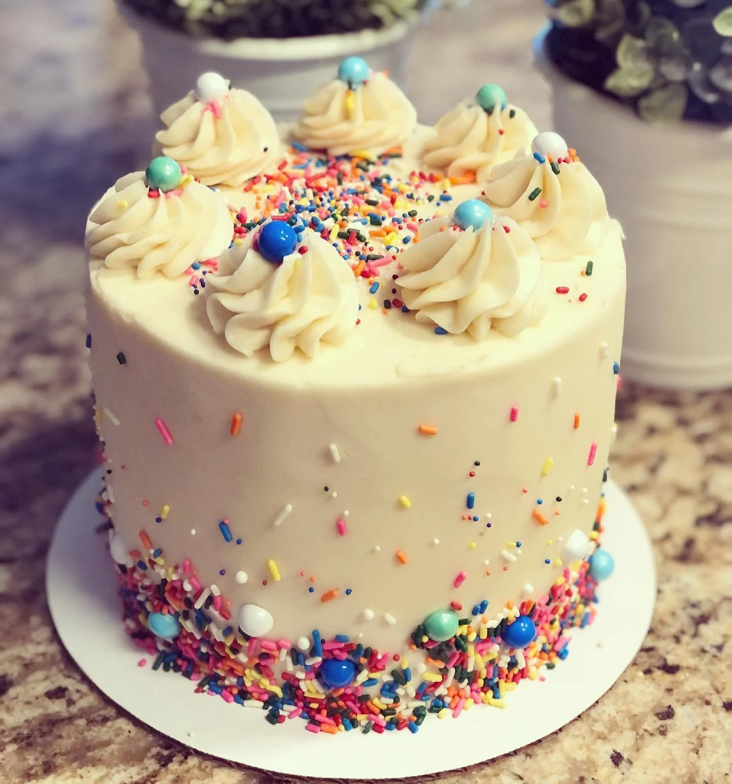 A white frosted birthday cake with colorful sprinkles and six dollops of white frosting topped with multi-colored round candies, on a white cake board on a speckled countertop.