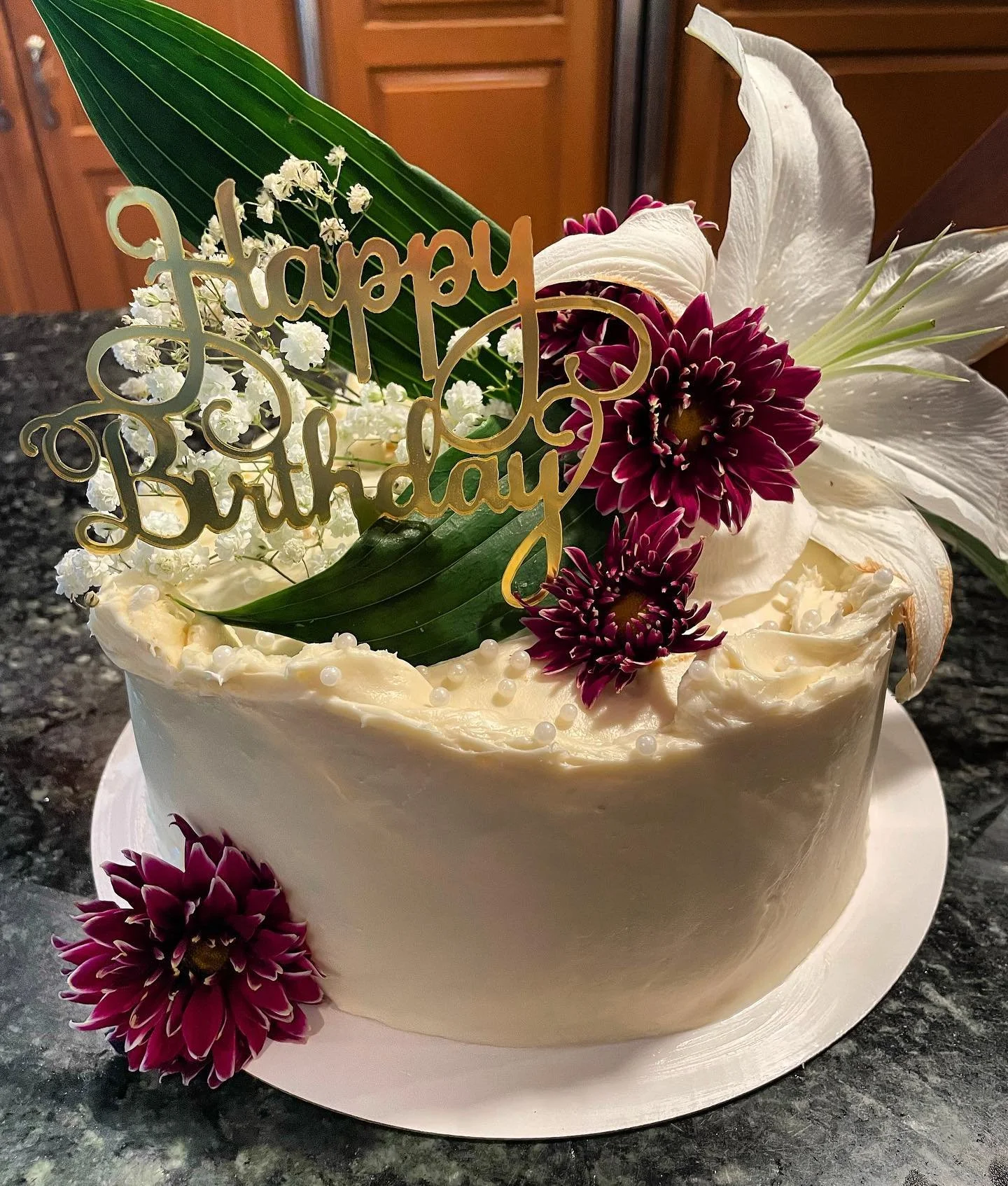 A birthday cake decorated with fresh flowers, including white lilies, burgundy chrysanthemums, and baby's breath, with a gold 'Happy Birthday' topper.