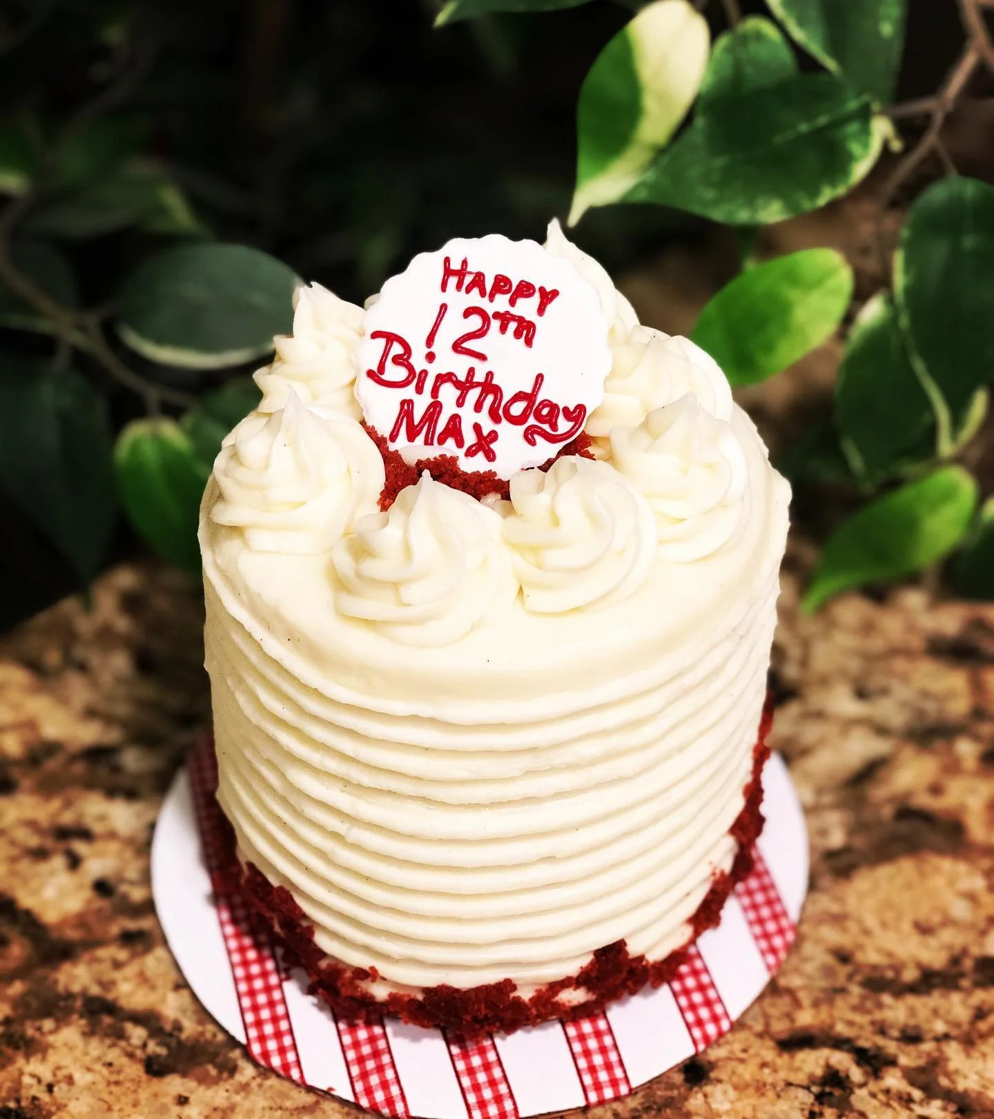 A birthday cake with white frosting and decorative piping on top, with a message written in red icing that says, "Happy 12th Birthday Max." The cake is on a red and white checkered napkin, and there are green leaves in the background.