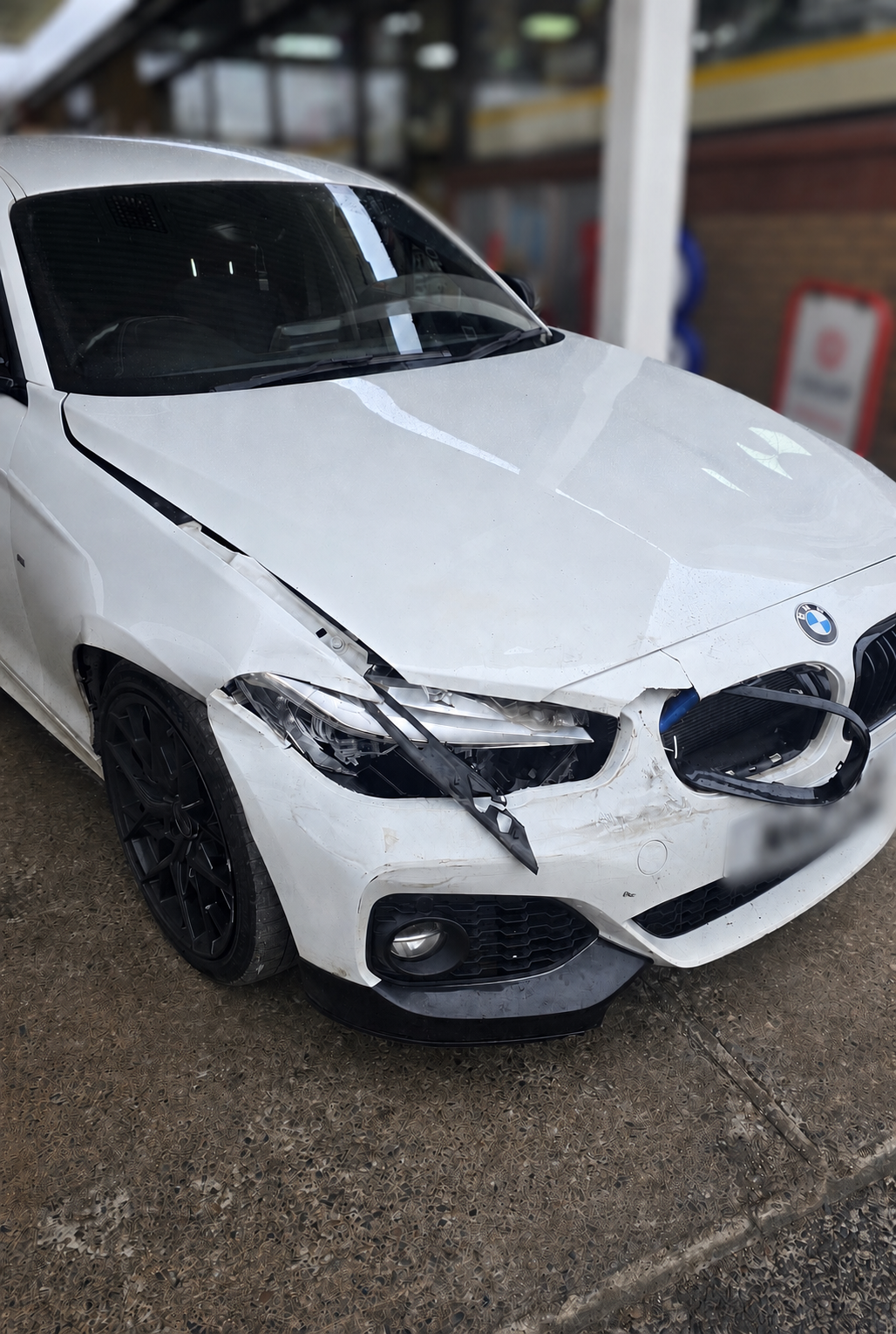 White BMW with front-end damage and missing right headlight, parked inside a garage or parking structure.