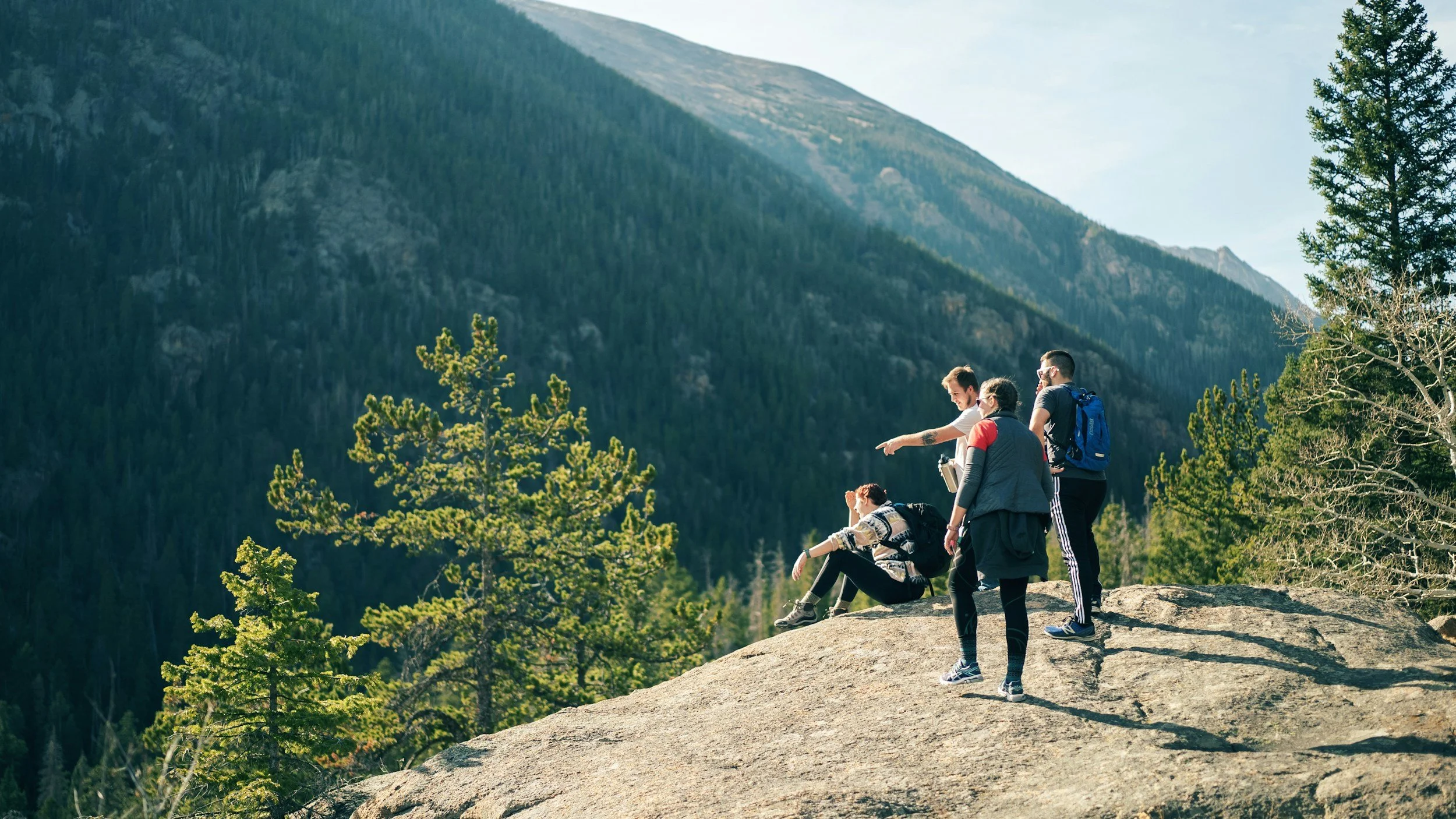 Group of five hikers standing on a rock ledge in a forested mountainous area, with some sitting and others standing, one pointing to the distance, surrounded by green trees and mountains in the background.