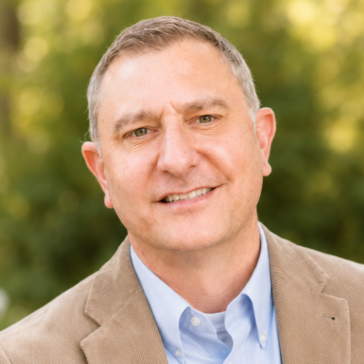 A middle-aged man with short gray hair, green eyes, and a warm smile, wearing a beige blazer and light blue collared shirt, standing outdoors with green foliage in the background.