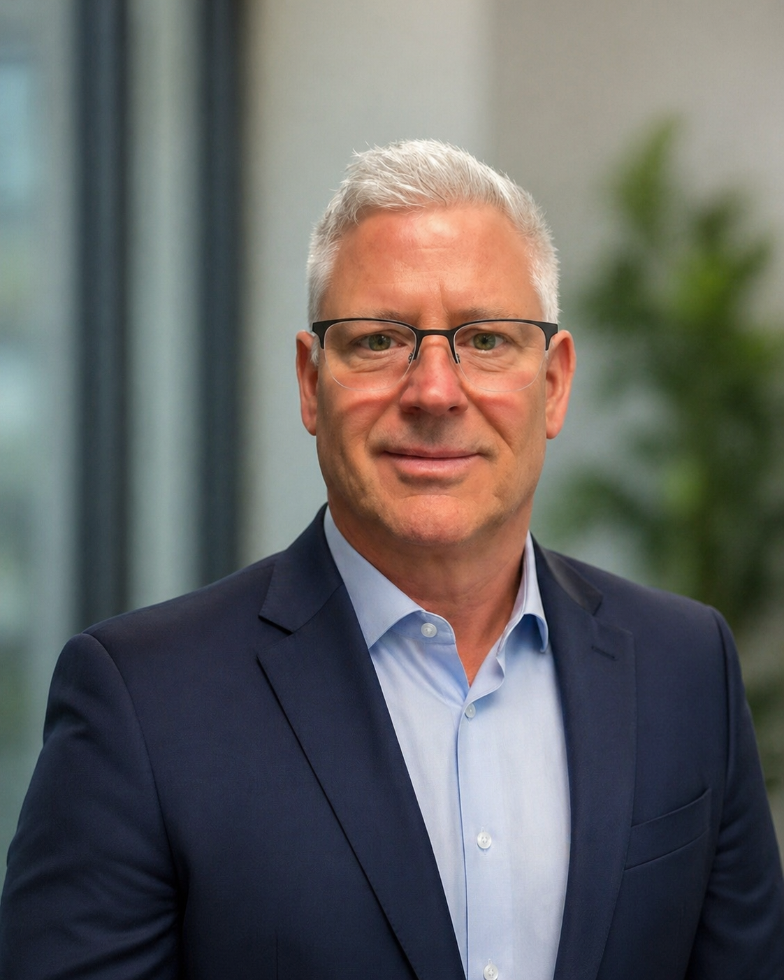 A middle-aged man with gray hair, wearing glasses, a dark blue suit, and a light blue shirt, standing indoors with a blurred background of office or building interior and a green plant.