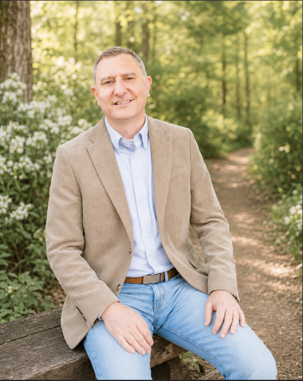 A man sitting on a wooden bench outdoors, wearing a tan blazer, light blue shirt, and jeans, with a background of green trees and sunlight filtering through the leaves.