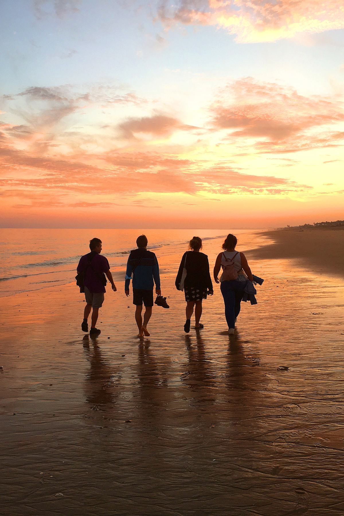 Four people walking along the beach at sunset with water reflecting the sky.