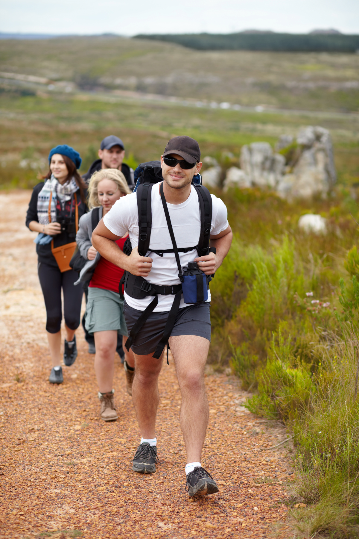 Group of four hikers walking along a dirt trail in a scenic outdoor landscape with rolling hills and rocks, dressed in casual hiking gear.