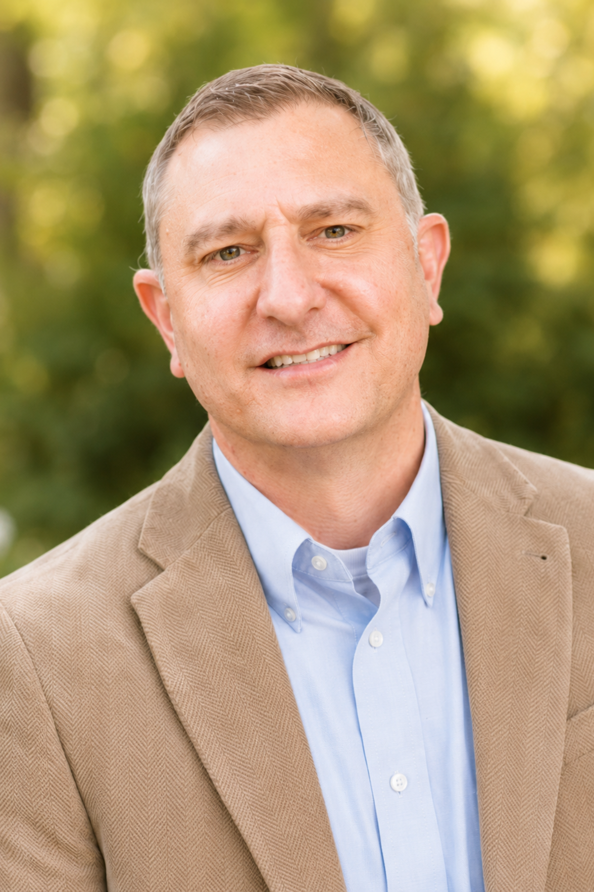 A portrait of a middle-aged man with short gray hair, wearing a light blue dress shirt and a tan blazer, outdoors with blurred green trees in the background.
