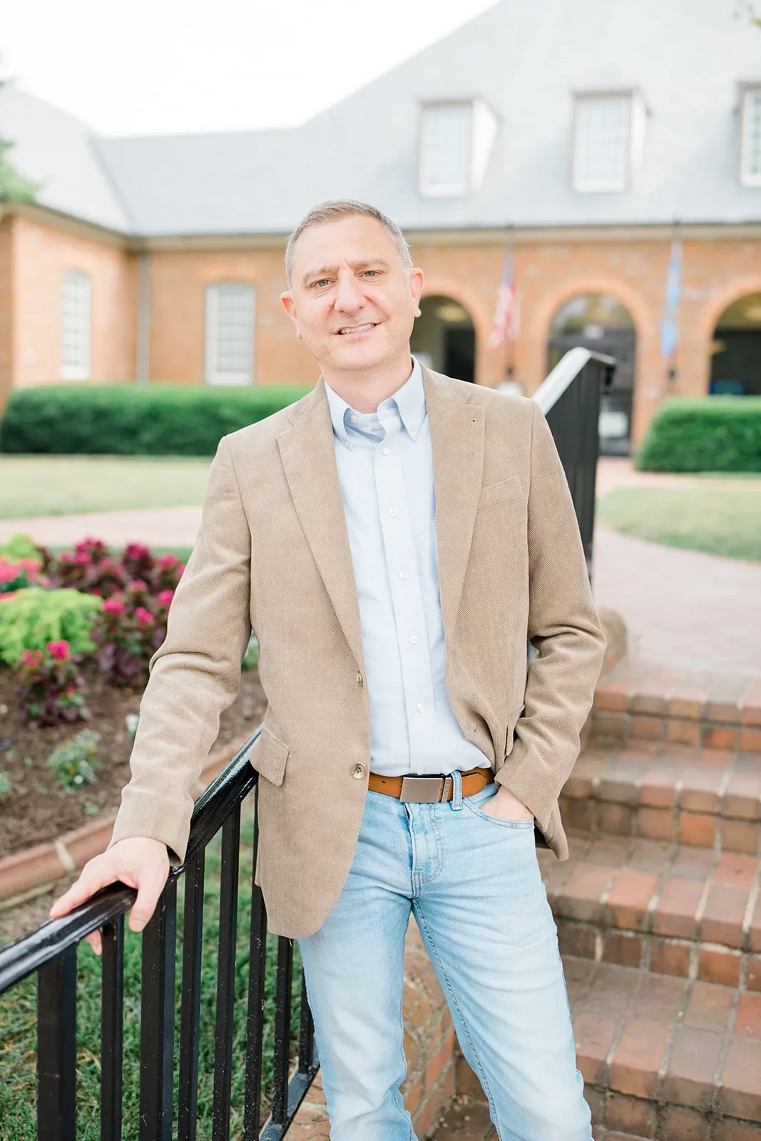 A man in a tan blazer and light blue jeans standing outdoors on brick stairs, holding onto a black metal railing, with a brick building with arched windows and flags in the background.