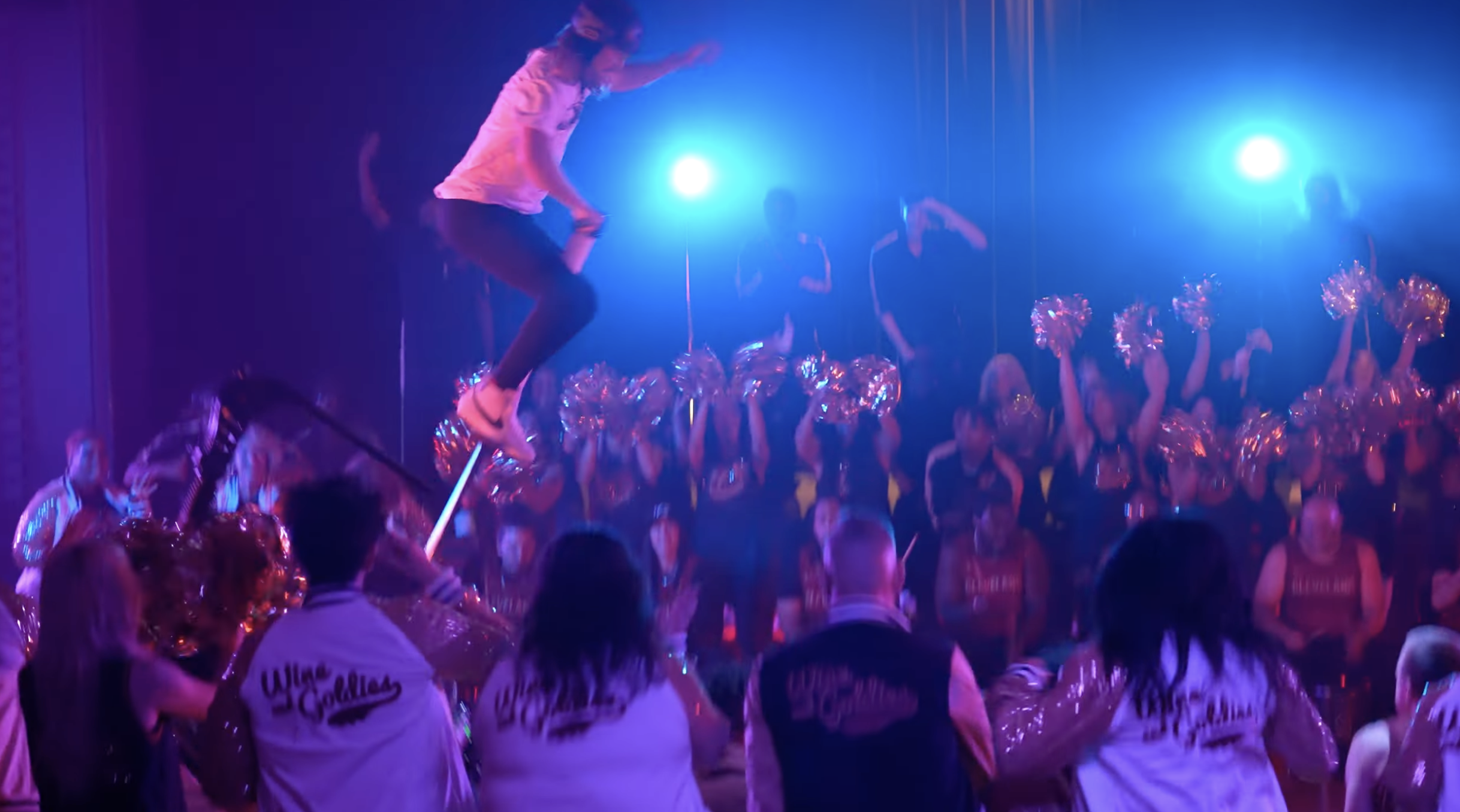 A person walking on a tightrope in a dark room illuminated by purple and blue lights, while a crowd of cheerleaders below watching and taking photos.