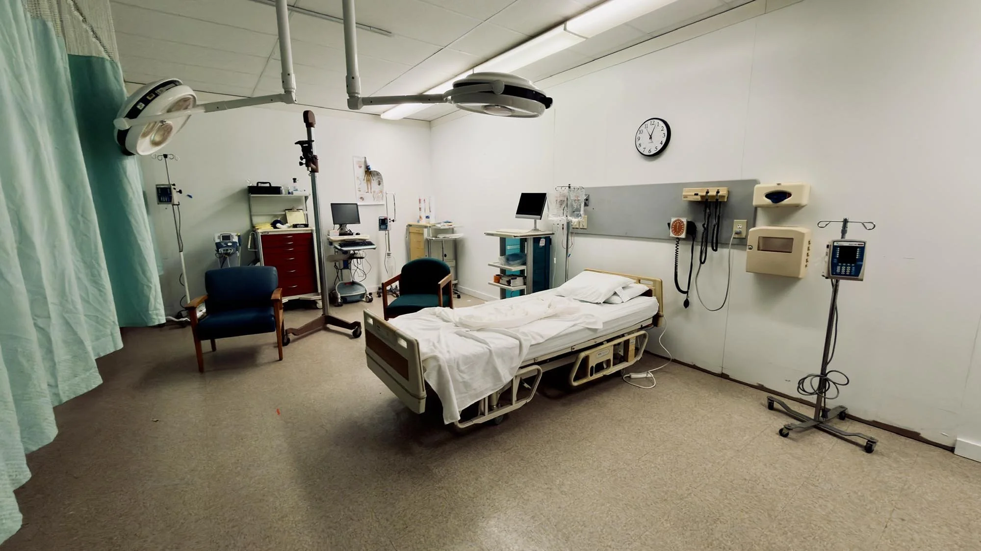 A hospital room with a bed, medical equipment, and a green privacy curtain on the left. The bed has white bedsheets and pillows. Various monitors and medical devices are attached to the wall and on stands, and two chairs are placed next to the bed. There is a wall clock showing 12:25.