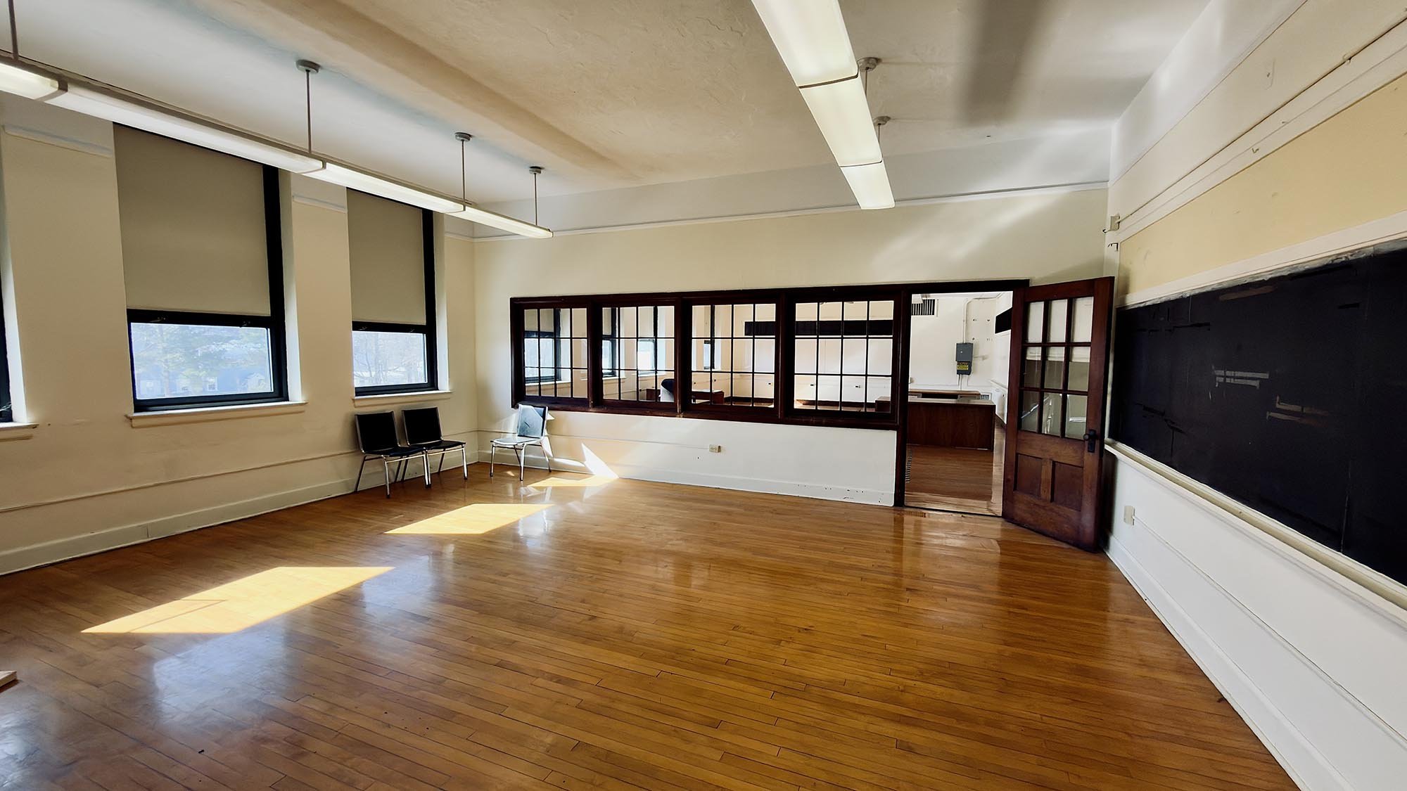 Empty room with wooden flooring, four windows letting in sunlight, chairs along the wall, and a blackboard with a wooden door leading to another room.