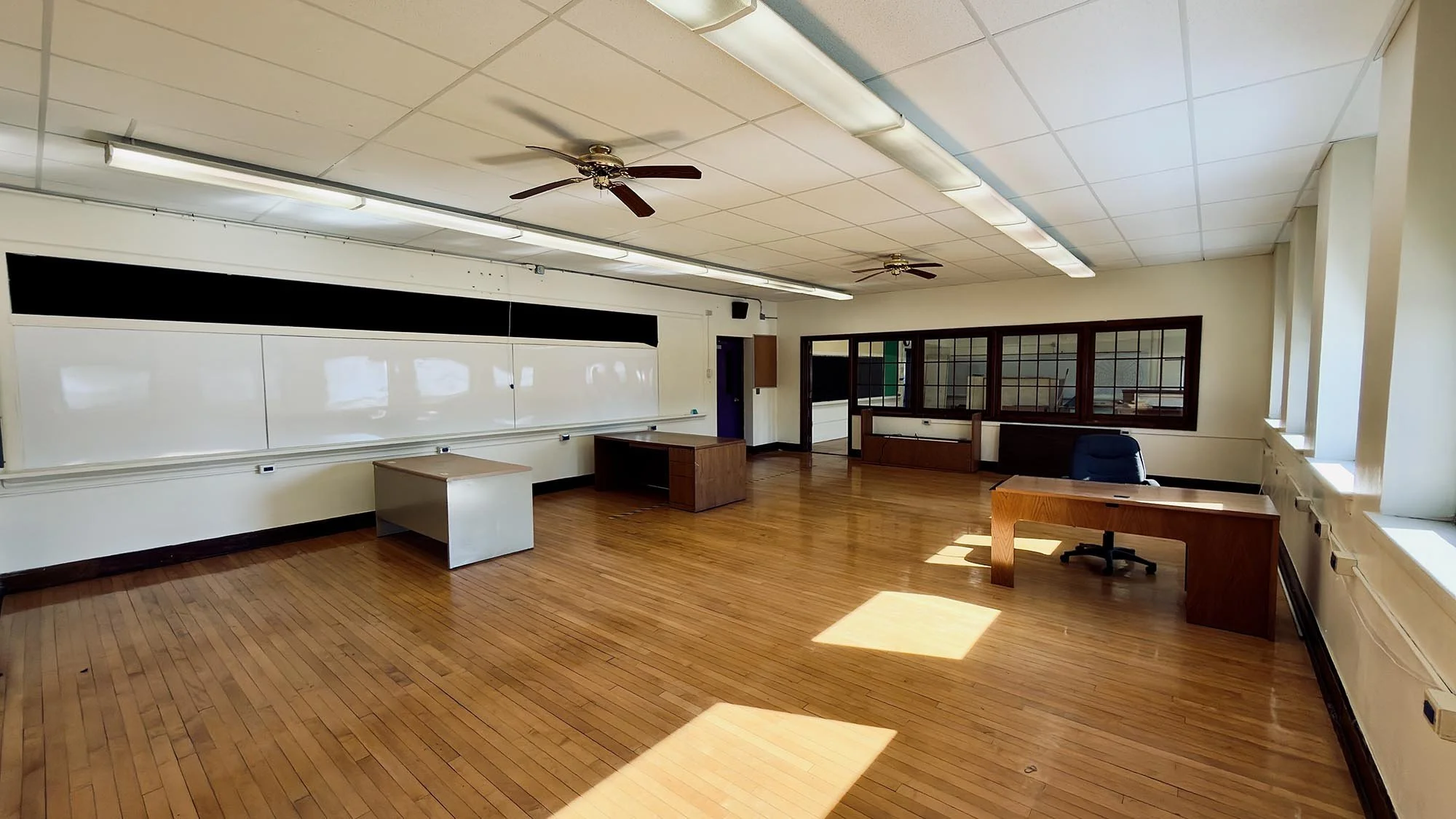 Empty classroom with wooden floors, desks, chairs, whiteboard, fluorescent lights, ceiling fans, and windows.