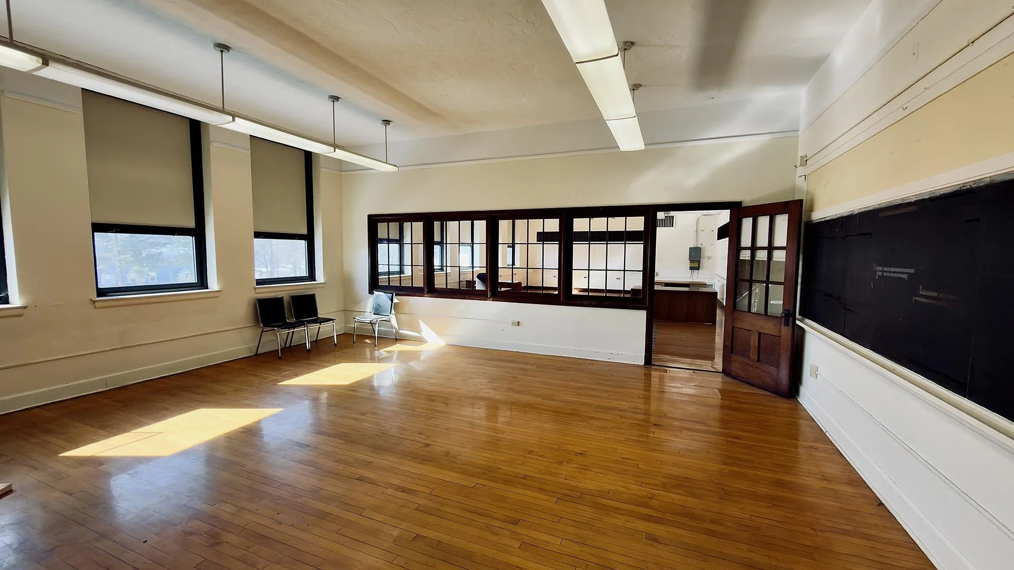 Empty classroom with three windows on the left, a blackboard on the right wall, wooden flooring, and a partitioned area with glass windows and a door in the back. Two chairs are placed near the windows.