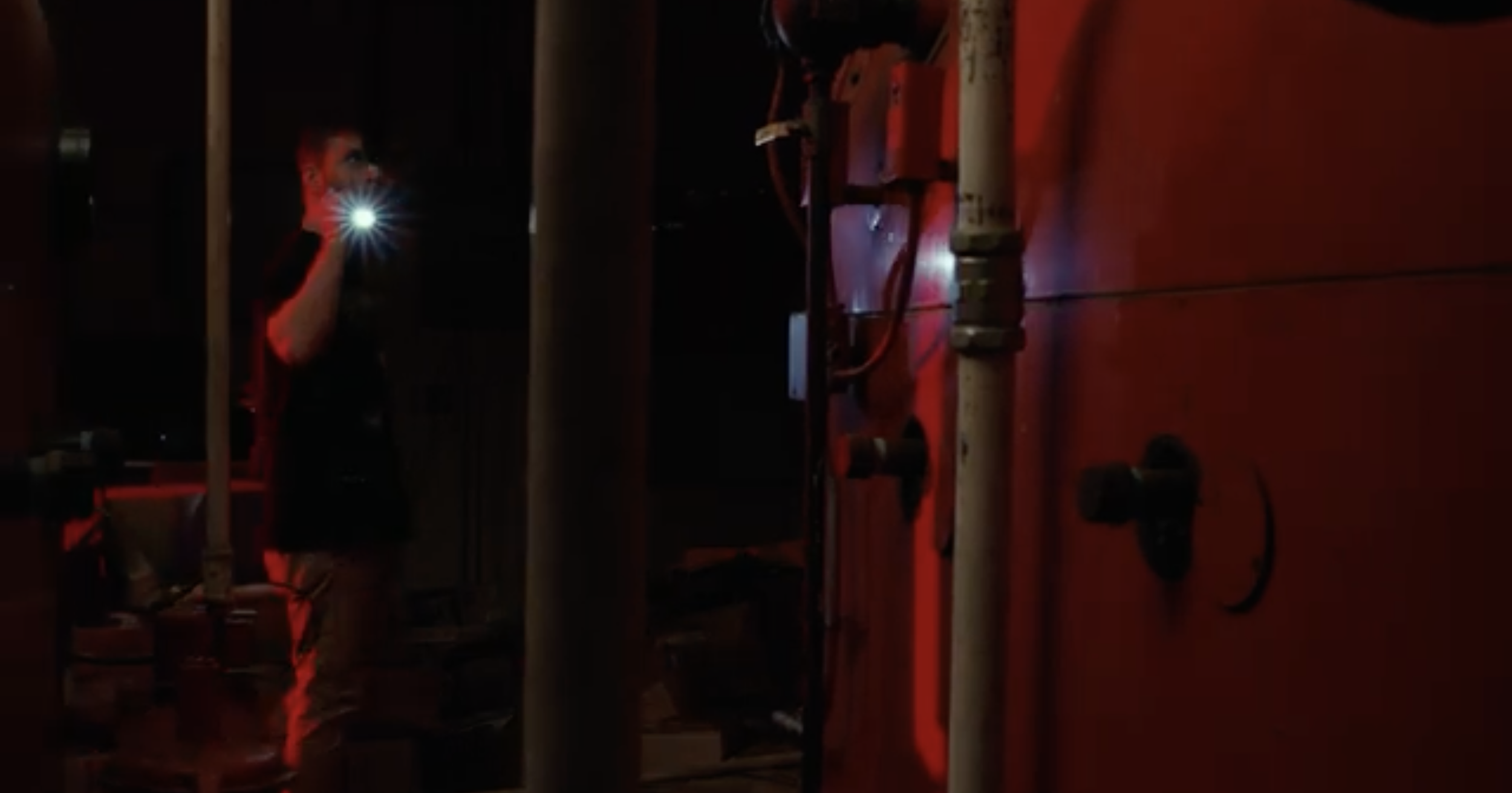 A person taking a photo or video with a phone flashlight in a workshop or garage with red lighting. There are tools and equipment on a red workbench and some machinery in the background.