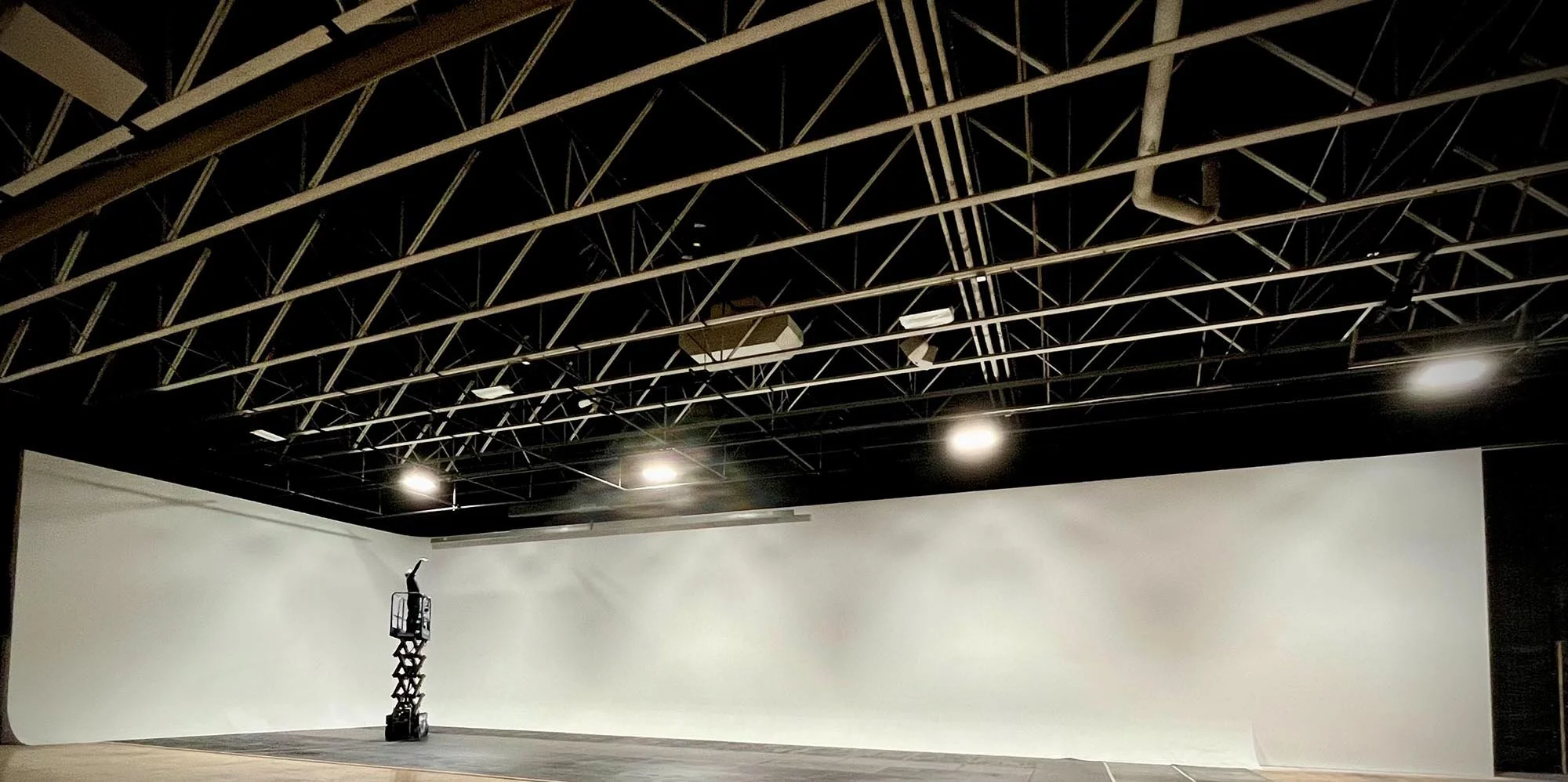 An empty theater or auditorium with a black ceiling featuring metal beams, ductwork, and bright overhead lights, and a large white screen on the front wall.