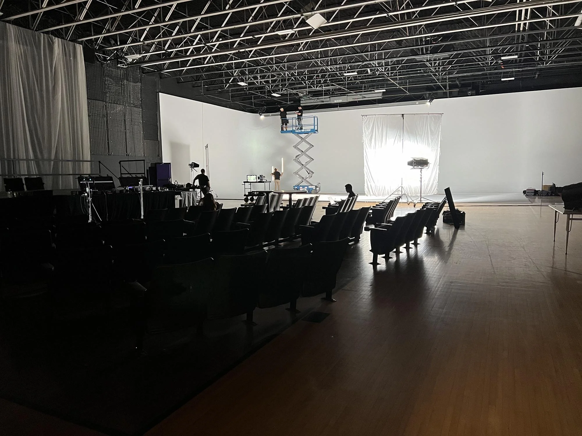 A large theater or studio with a white screen or backdrop, with people preparing the space using a scissor lift and equipment. Rows of empty chairs are visible in the foreground.