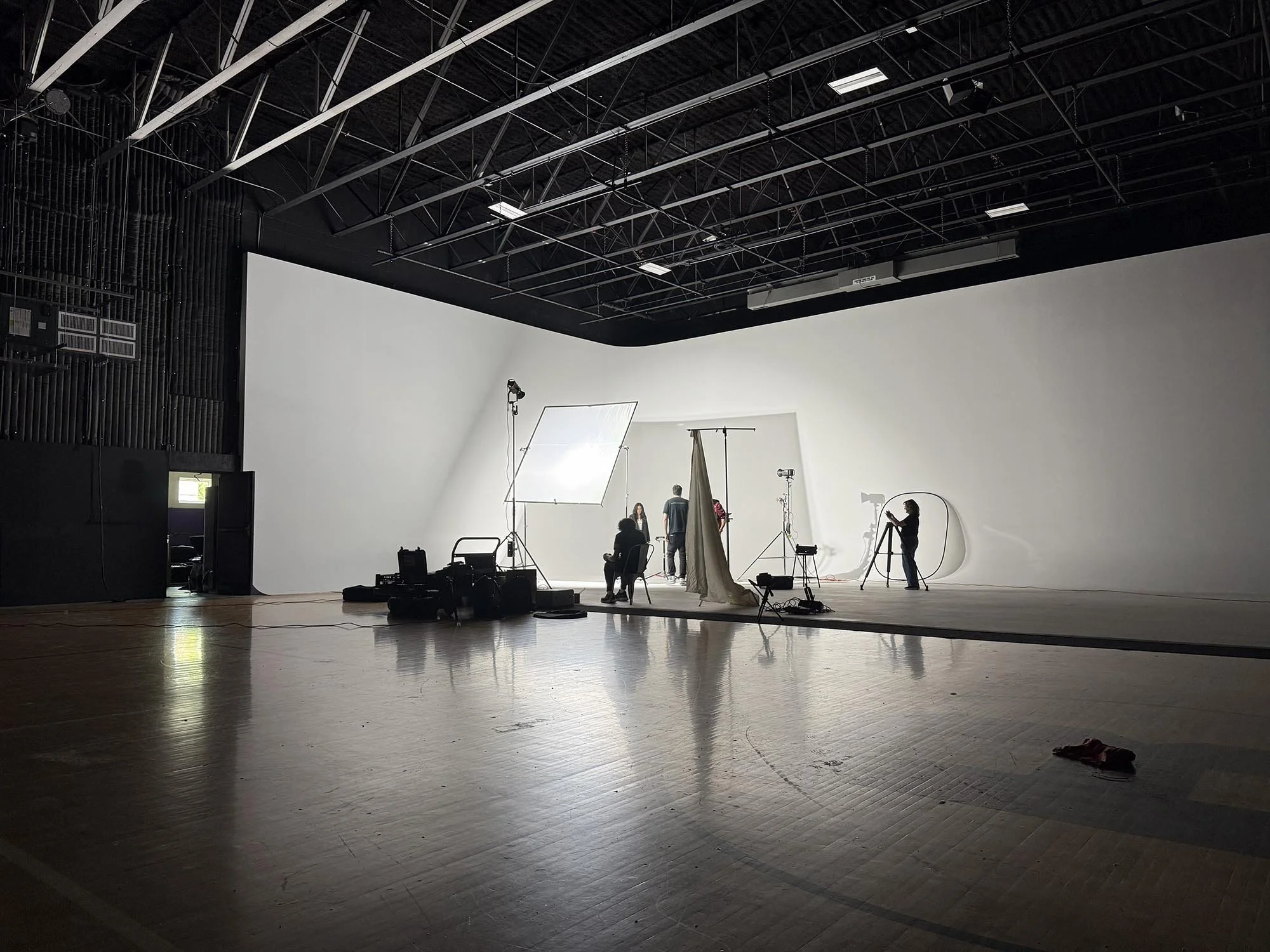 A photography studio or film set with a large white backdrop, studio lighting equipment, and people working around the set, some sitting and others standing.