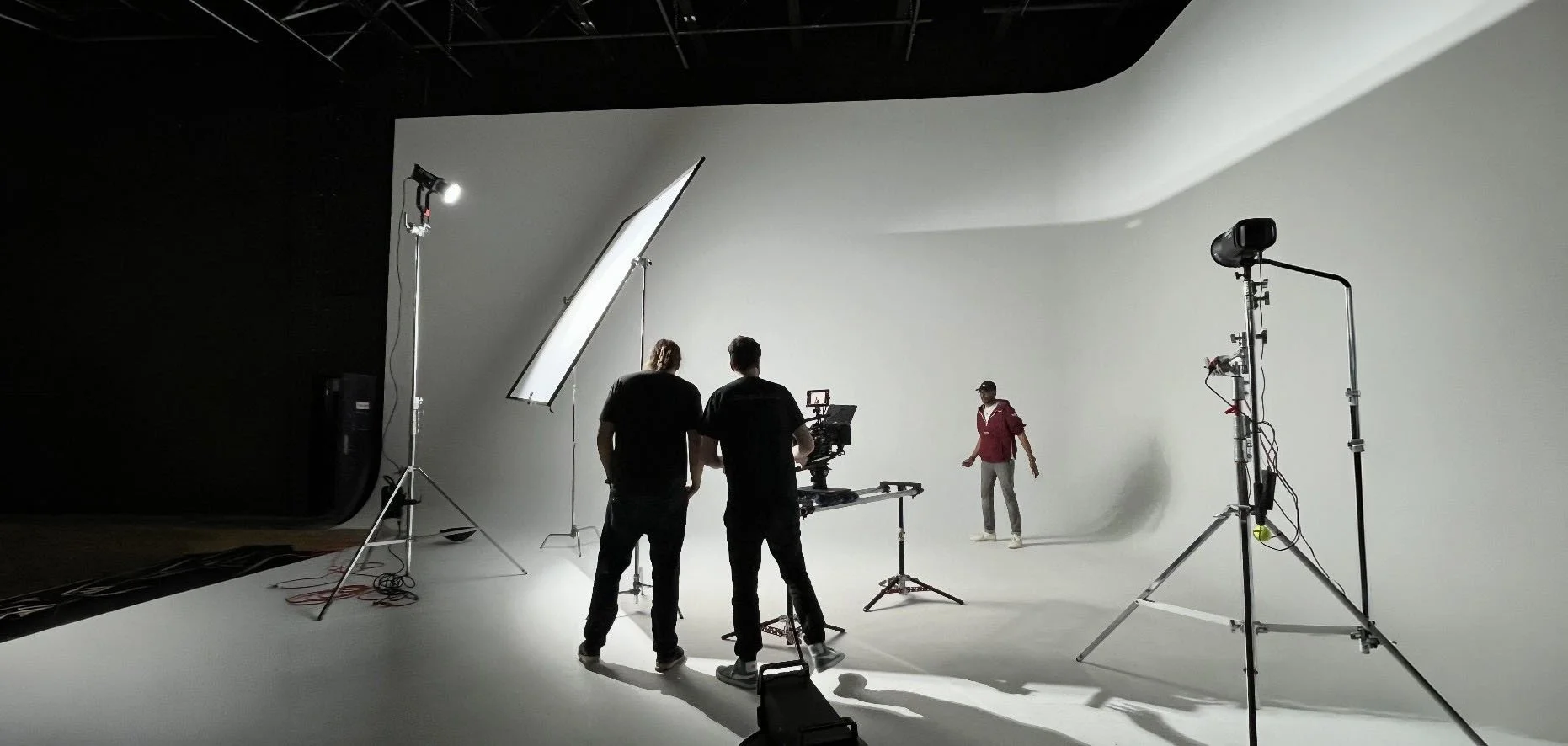 Studio set with a white seamless backdrop, featuring professional lighting equipment, including softboxes and reflectors, while two crew members operate a camera and a third person stands in the background.
