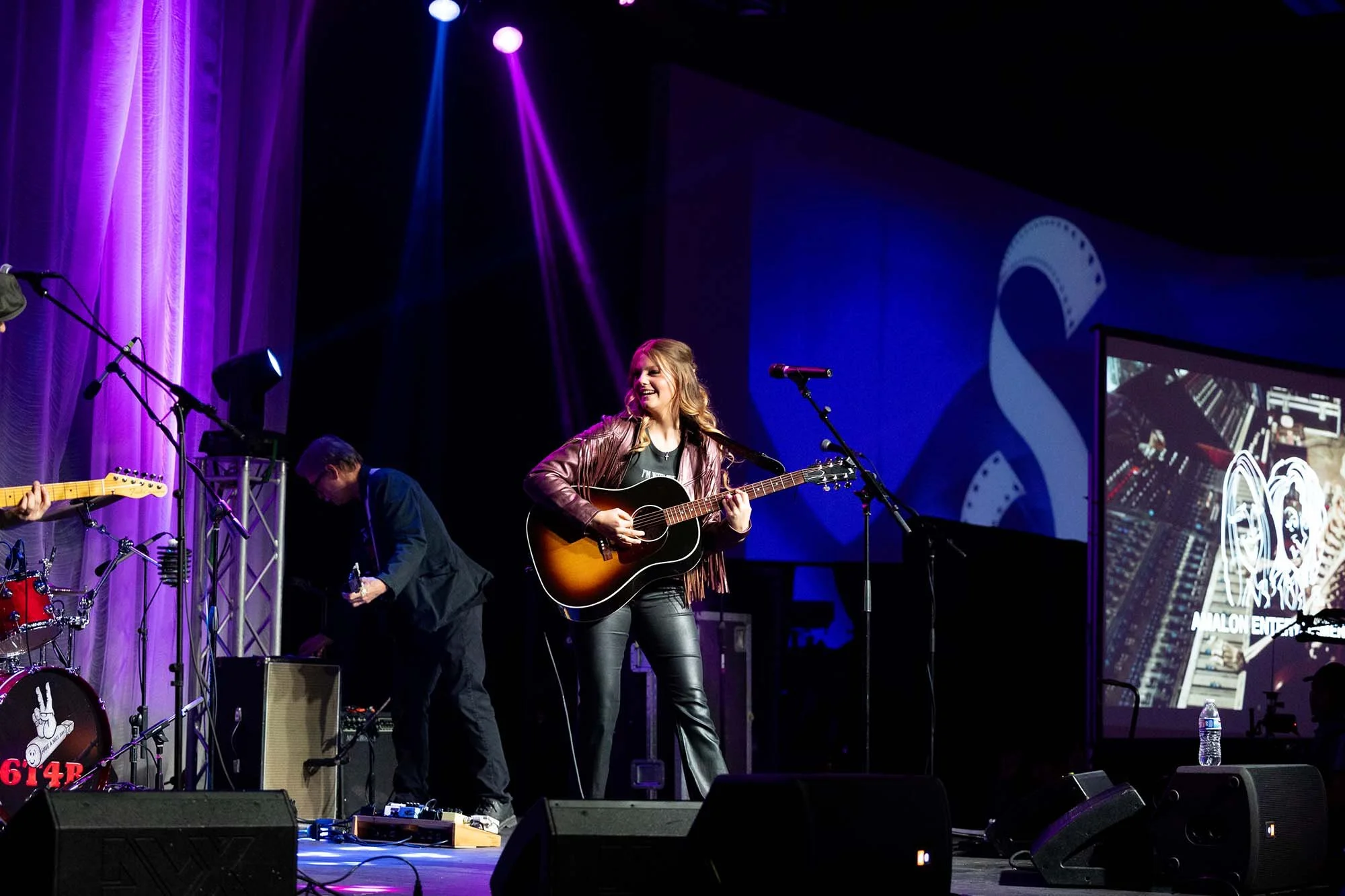 A woman with blond hair playing an acoustic guitar on stage during a live performance, with a purple curtain backdrop and purple spotlight, accompanied by band members under stage lighting.