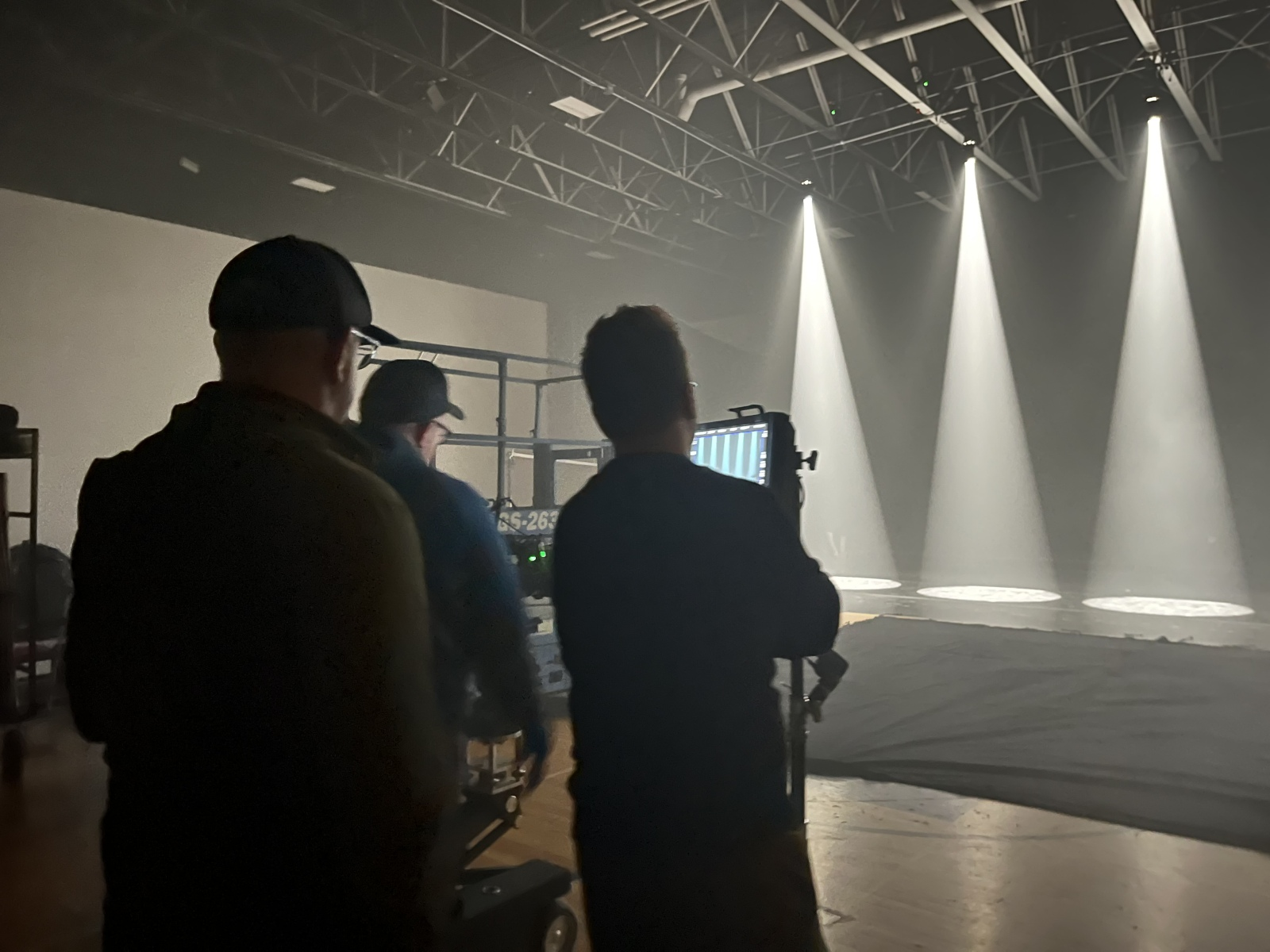 Three crew members working behind the scenes on a stage with bright overhead lights, preparing for a performance or filming.