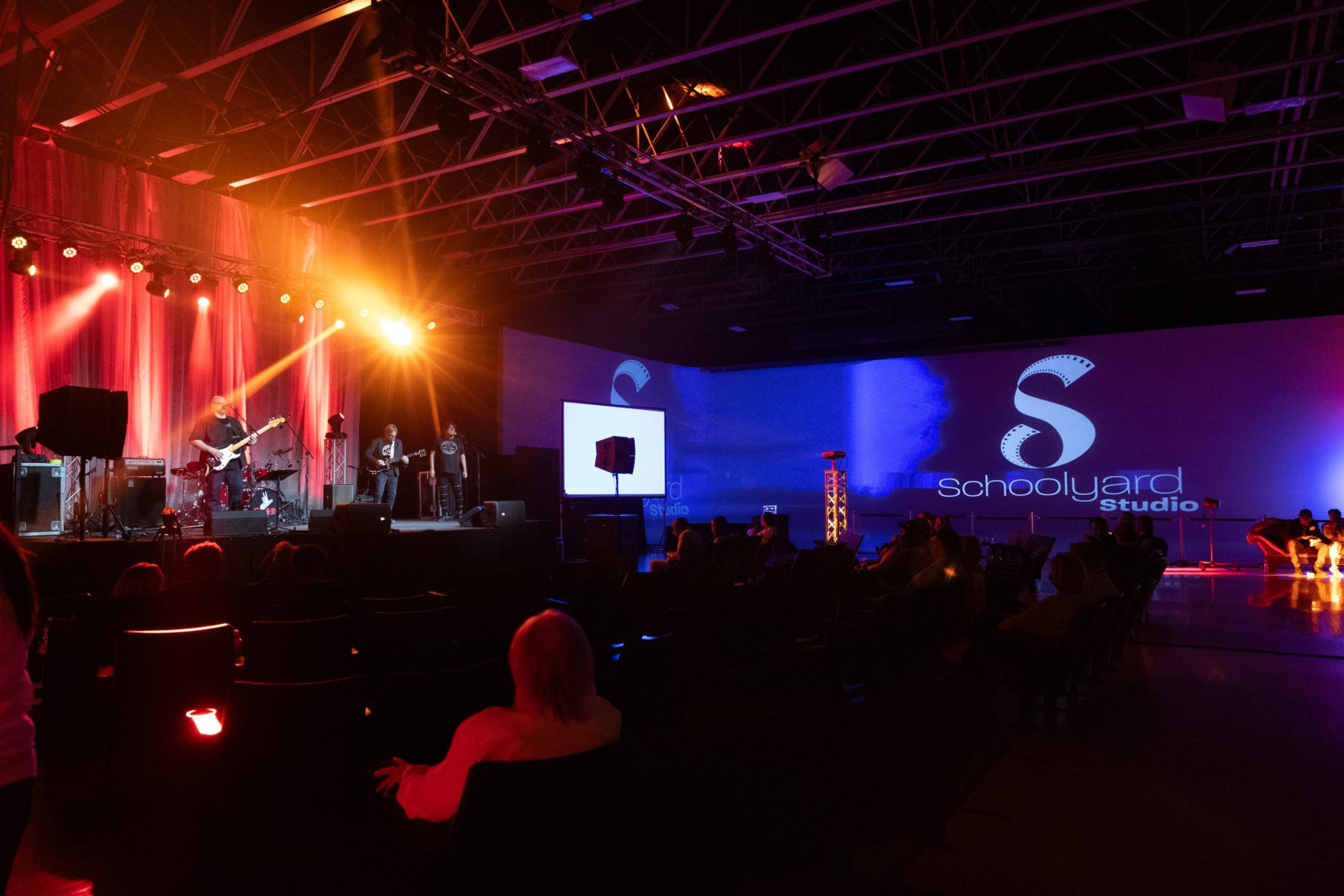 Music band performing on stage at Schoolyard Studio with colorful lighting and audience in seats.