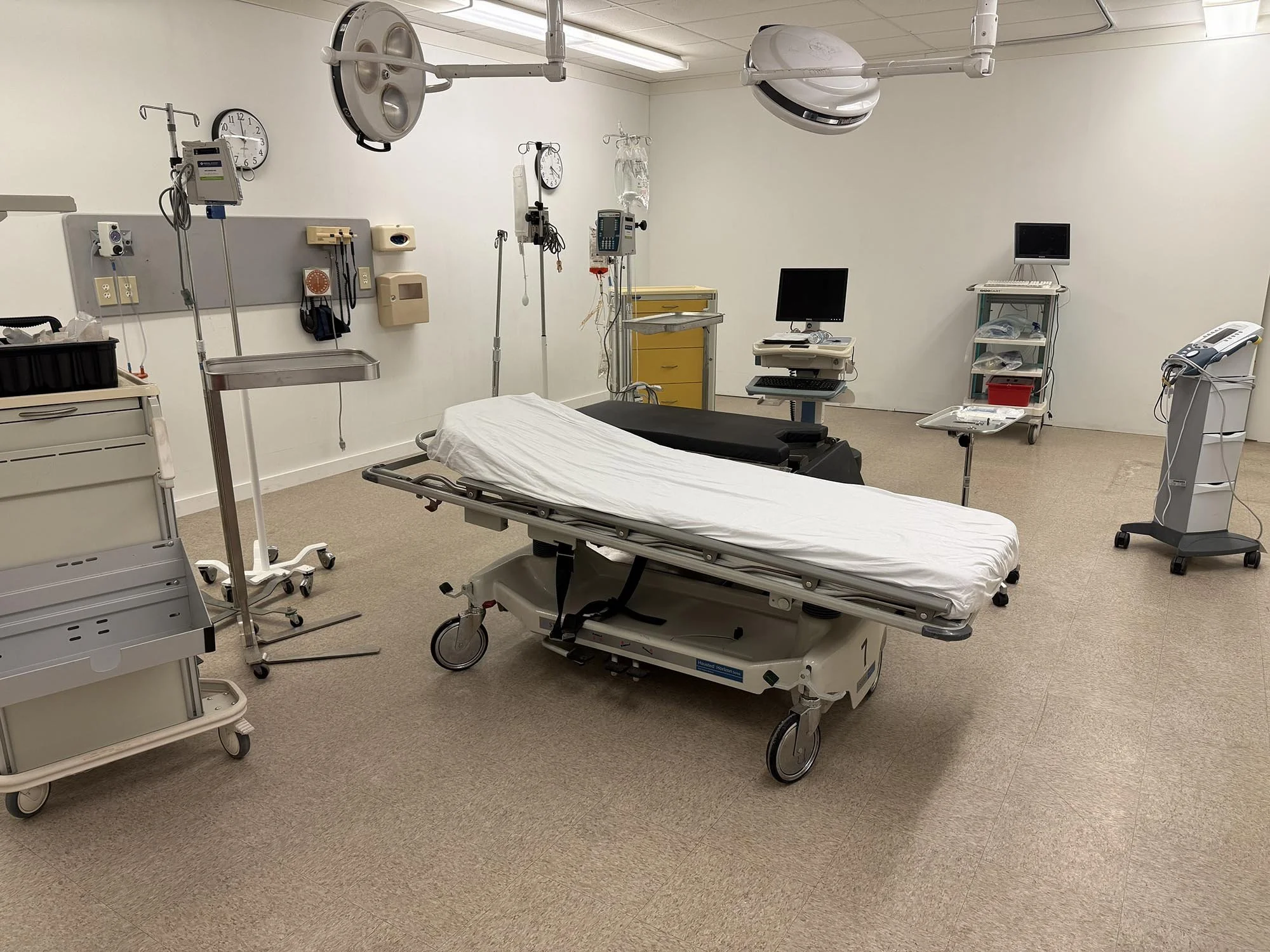 Empty hospital room with a hospital bed, medical equipment, monitors, and overhead surgical lights.