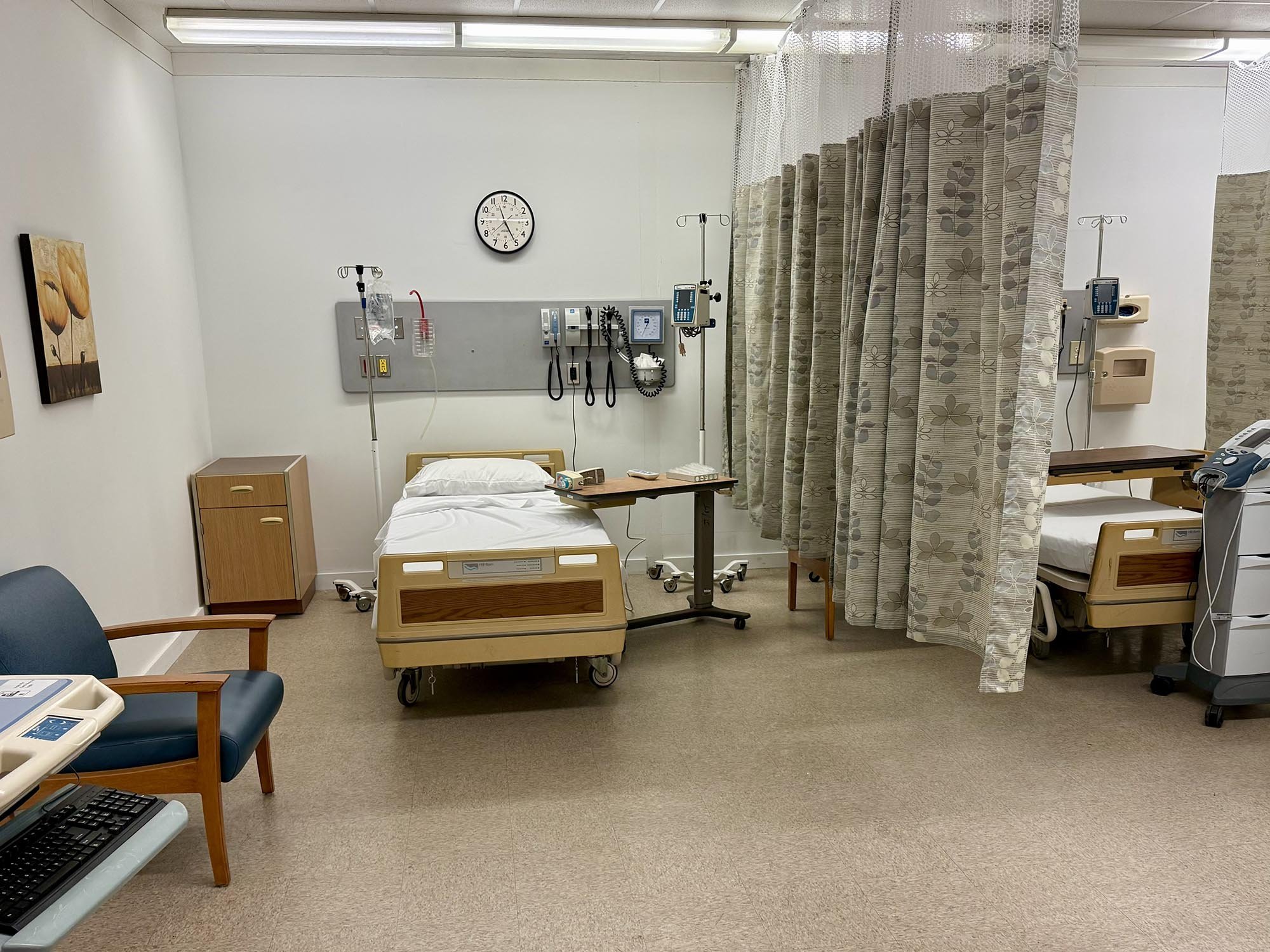 A hospital room with two hospital beds separated by a curtain. Each bed has medical equipment and IV stands. There are chairs and a small wooden cabinet nearby, a clock on the wall showing 10:09, and some wall-mounted medical devices.