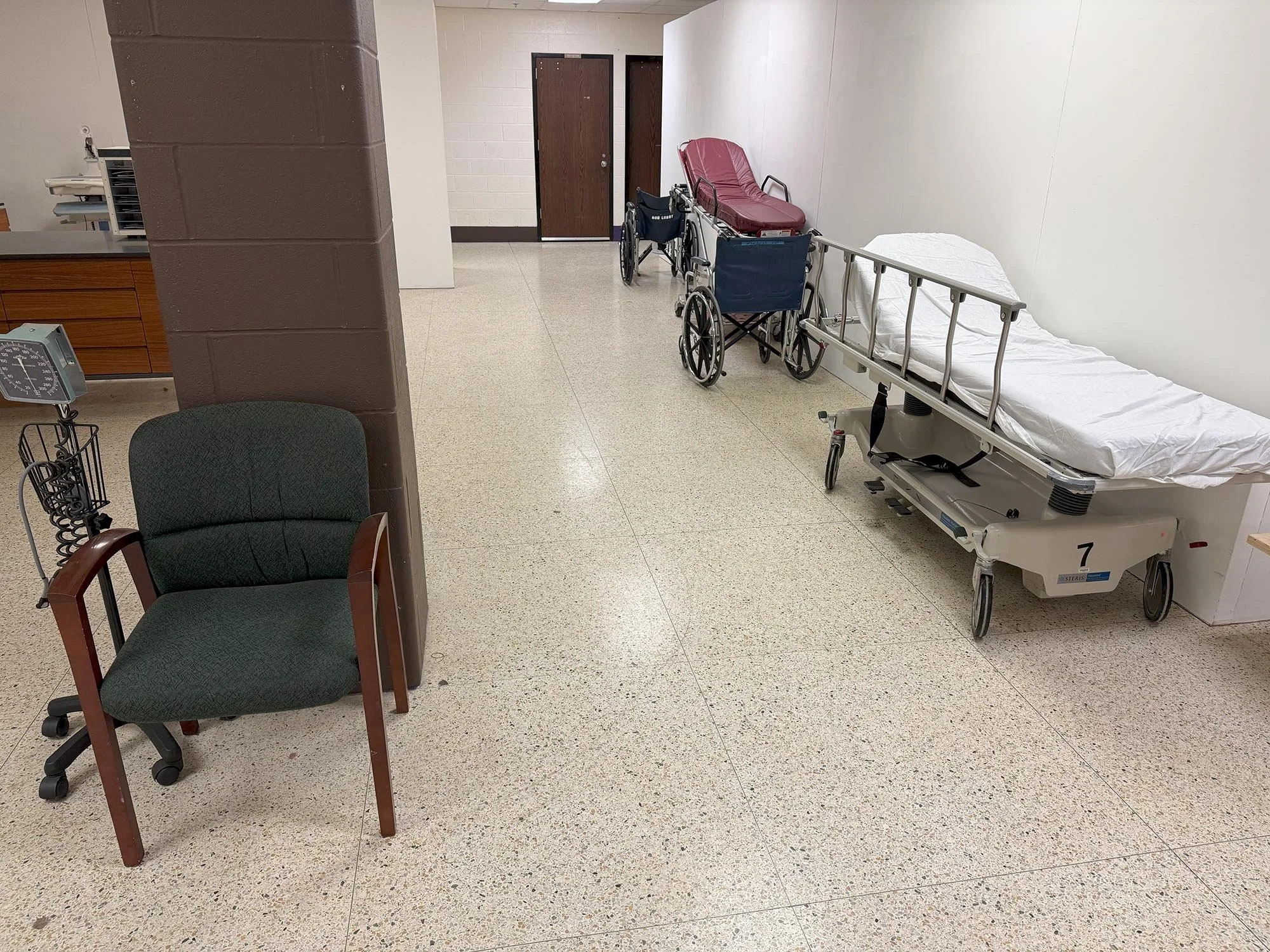 An empty hospital room with a hospital bed, a wheelchair, and a burgundy recliner chair. The room has beige tiled floors, white walls, and two closed brown doors.