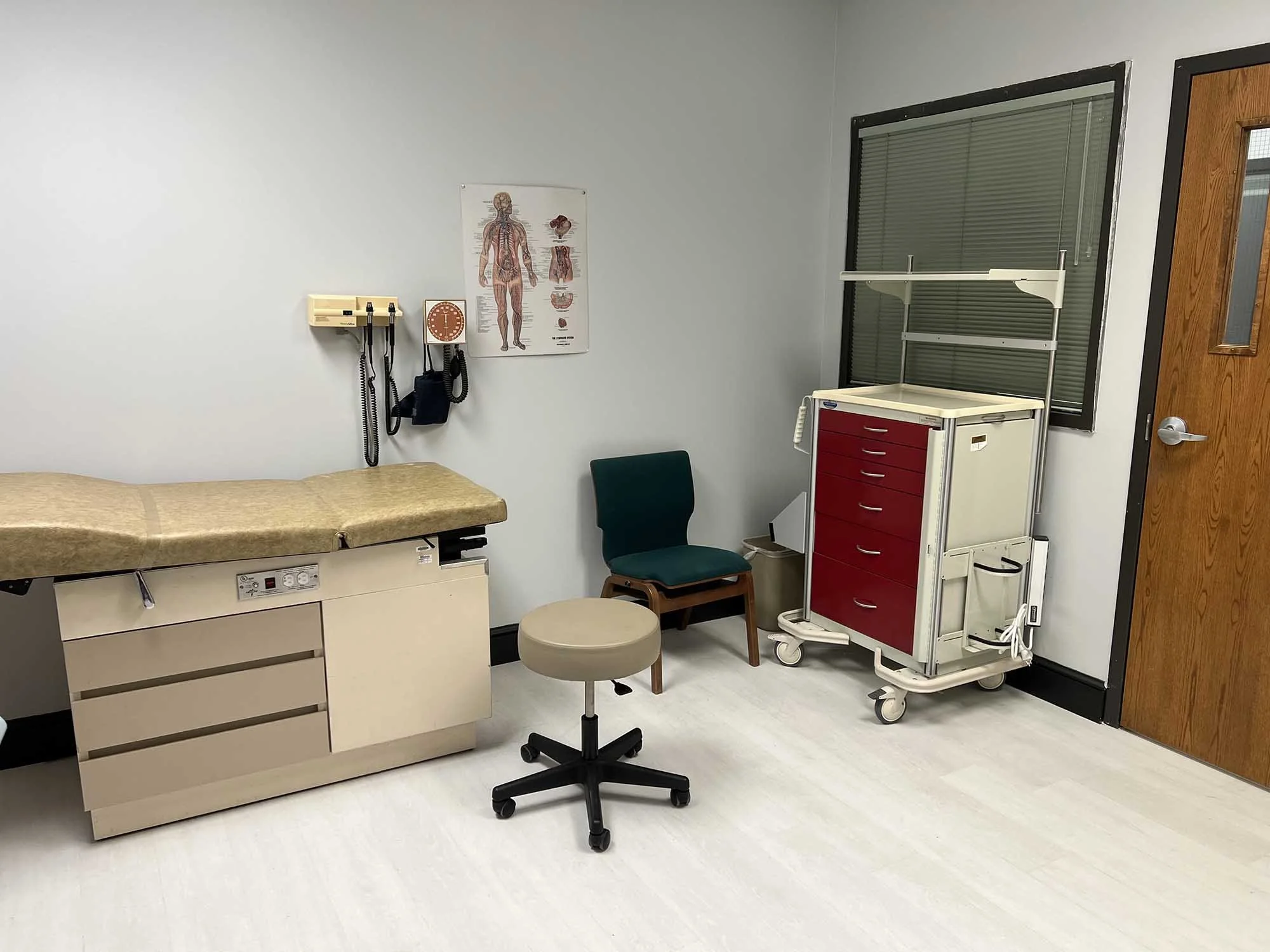 A hospital examination room with an examination table, a chair, a stool, medical posters, a wall-mounted blood pressure cuff, and a medical cart.