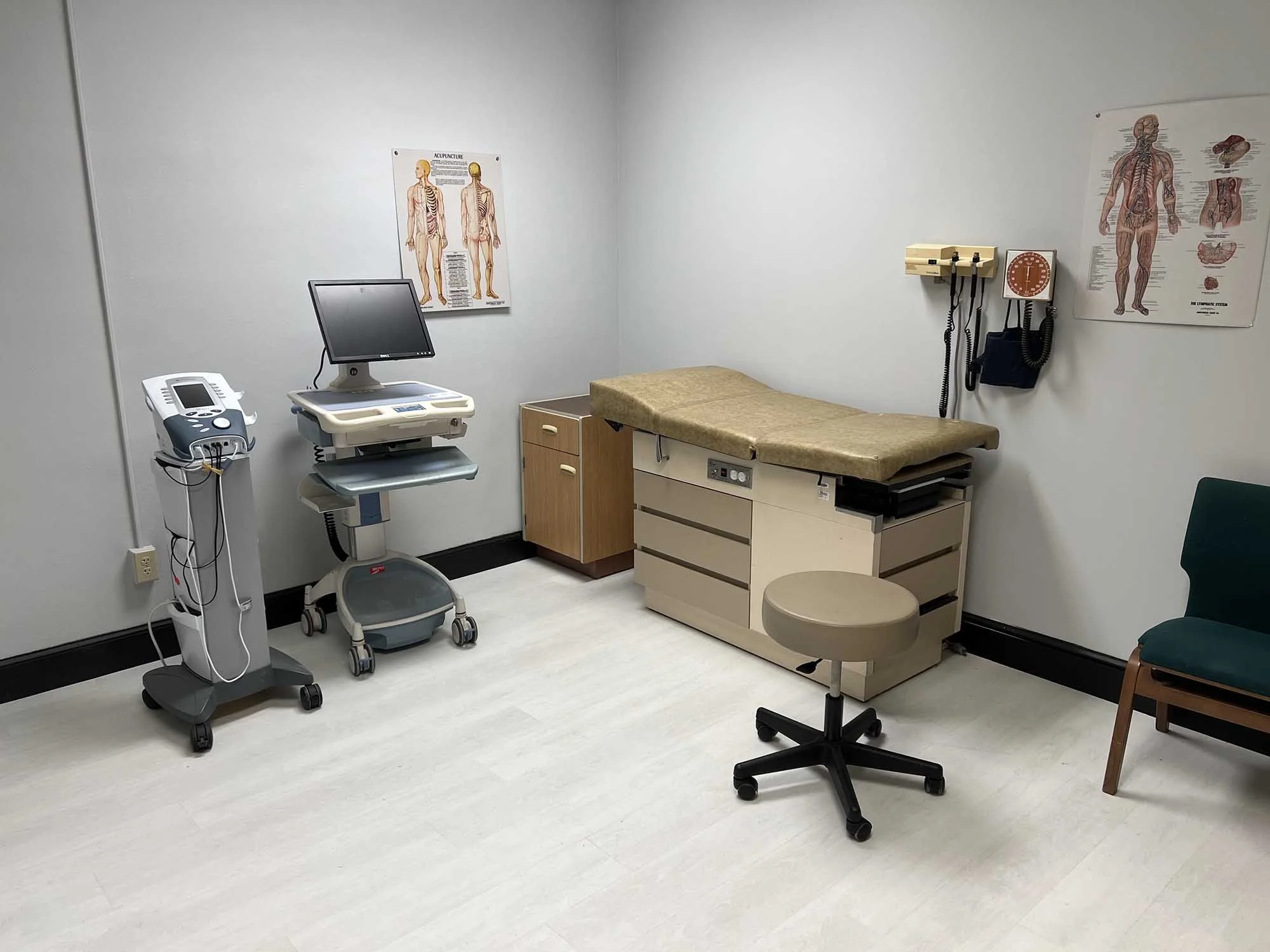 Empty medical examination room with an examination table, an ergonomic stool, medical posters on anatomy and acupuncture, a monitor, medical equipment on a cart, a wall-mounted blood pressure monitor, and a green chair.