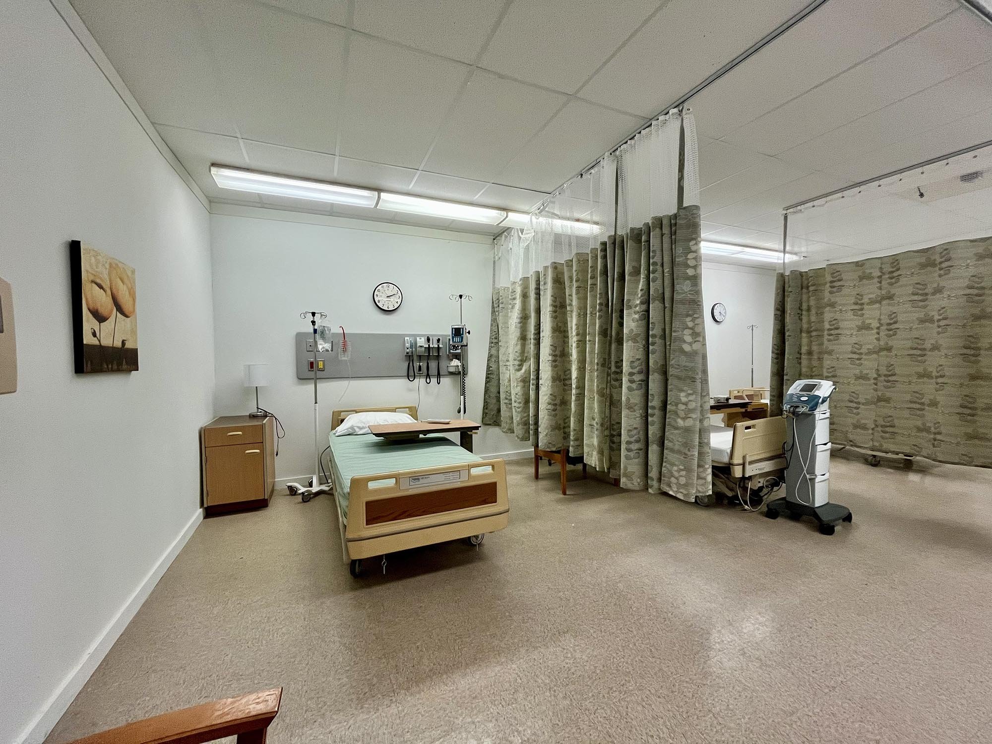 A hospital room with an empty bed, medical equipment, privacy curtains, a bedside table, and a clock on the wall.
