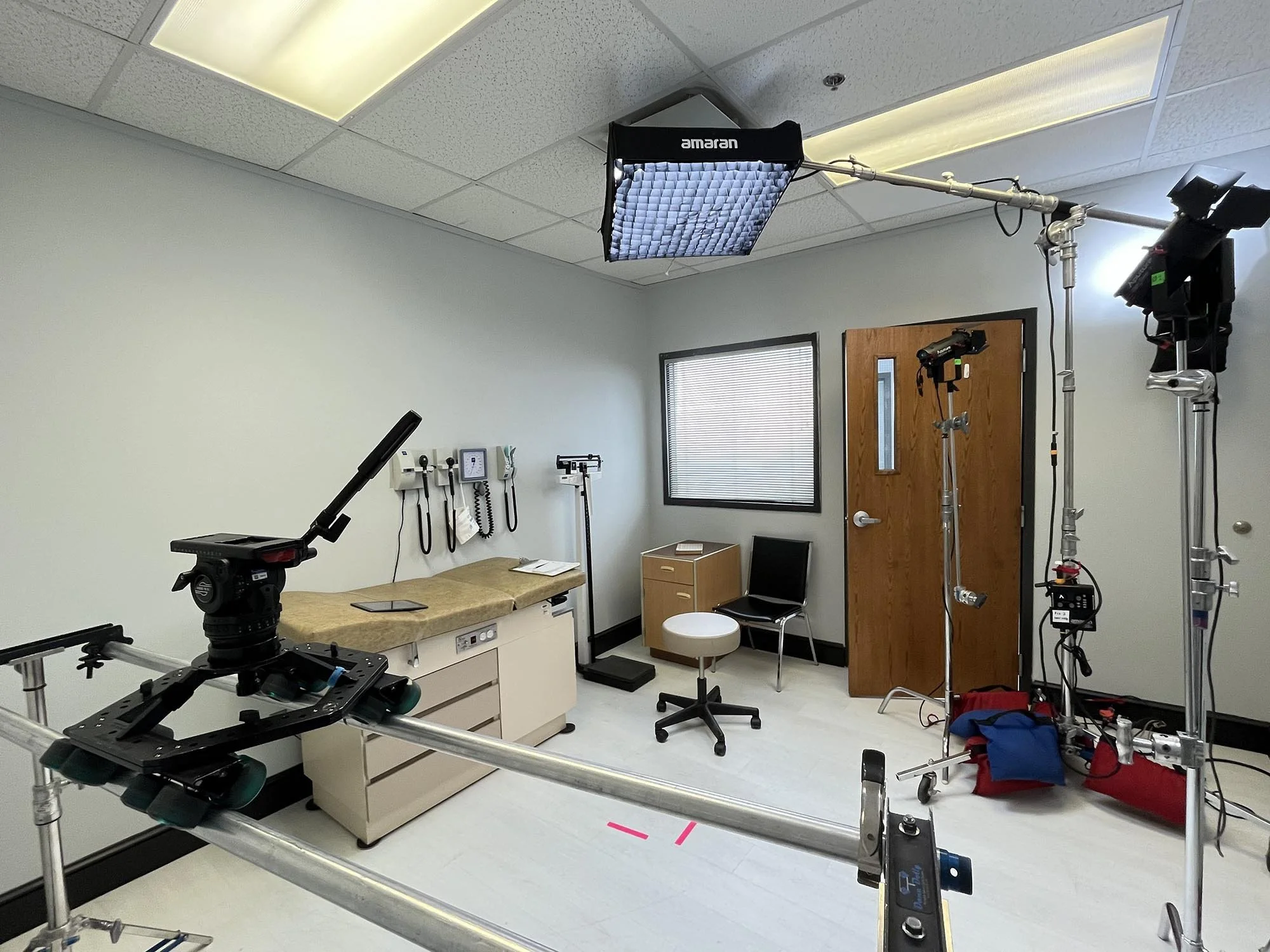 Film or television production set with lighting equipment, camera on a stabilizer, examination room furniture, medical tools on the wall, and a closed wooden door.
