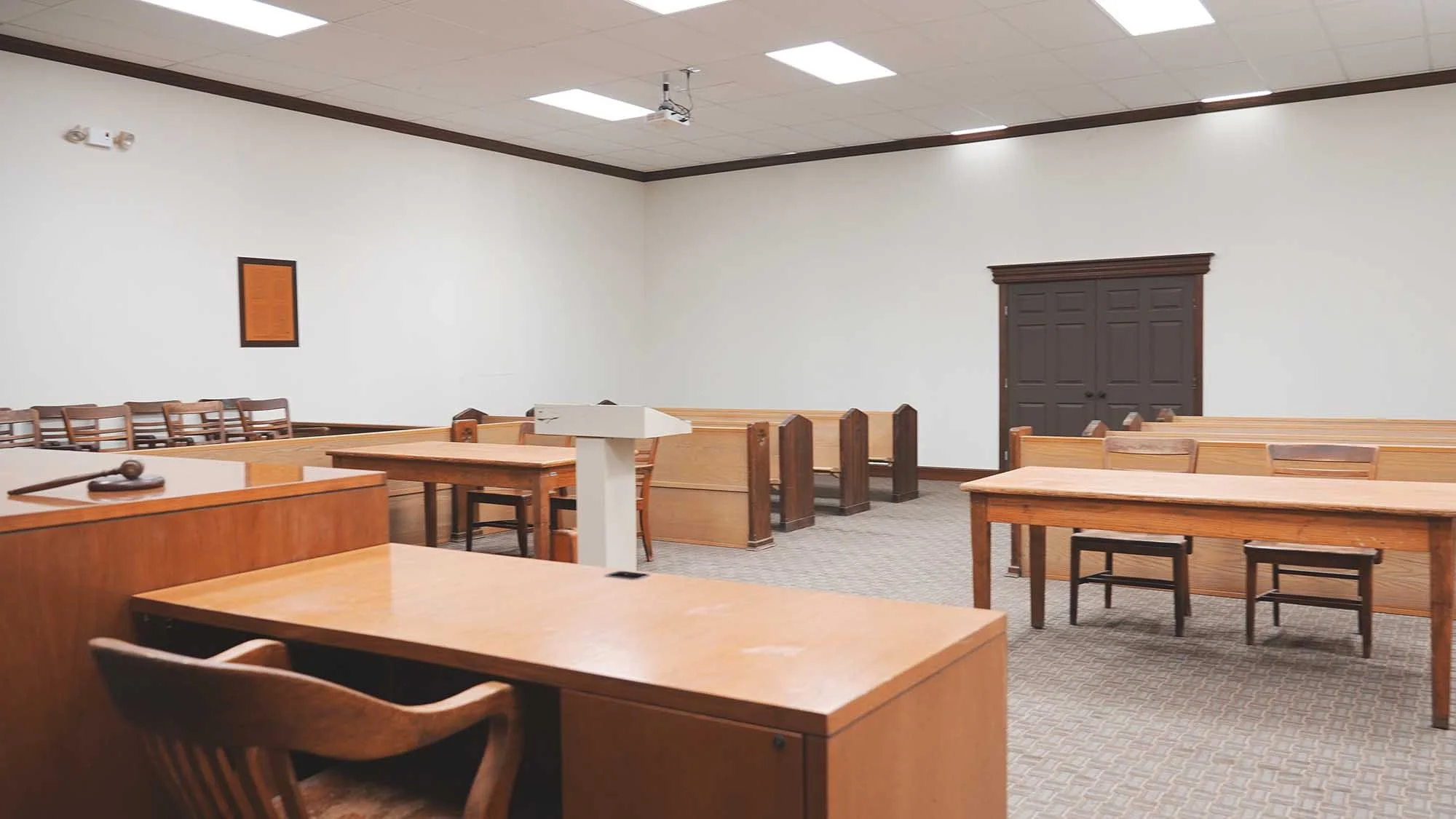 A courtroom with wooden tables, chairs, and a judge's bench. The room has a drop ceiling with fluorescent lighting and a projector mounted on the ceiling. There's a dark wooden double door on the back wall and a framed document or picture on the wall