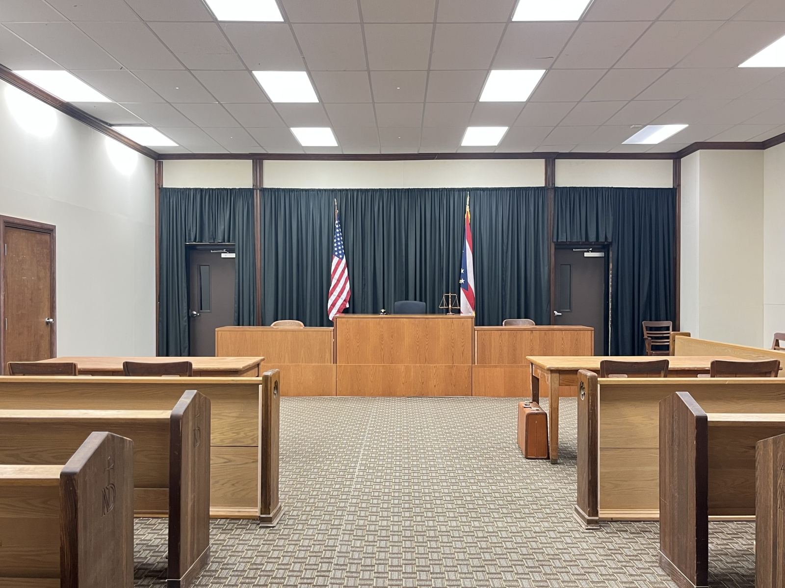 An empty courtroom with wooden benches, a judge's bench, and two flags, one American and one Missouri state, behind the judge's bench.