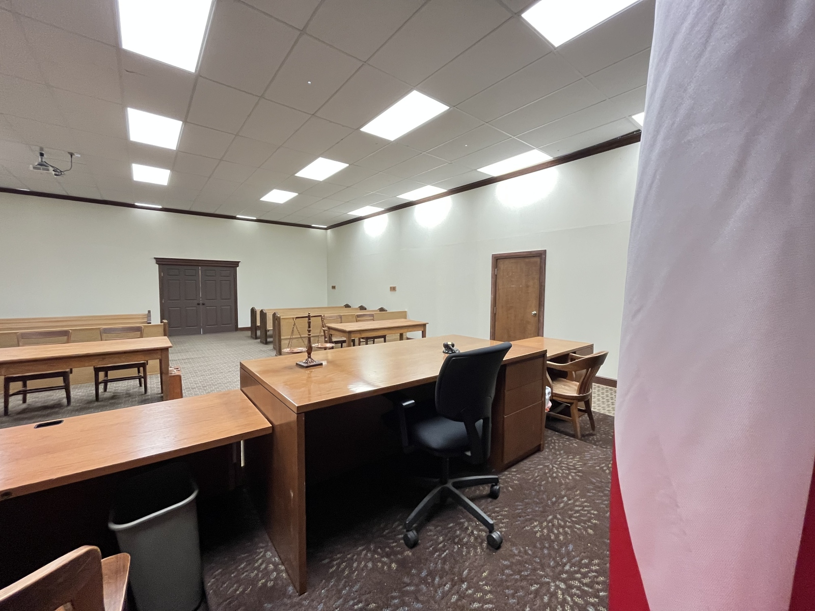 Empty courtroom with wooden benches, judge's table, and a flag partially visible on the right side.