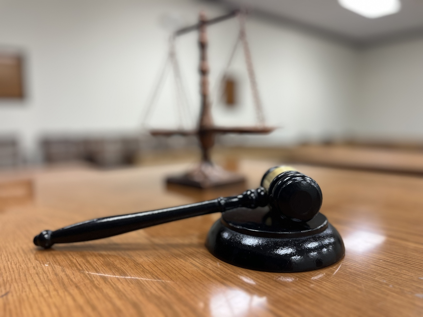 Close-up of a judge's gavel resting on a wooden table in a courtroom with a blurred scale of justice in the background.