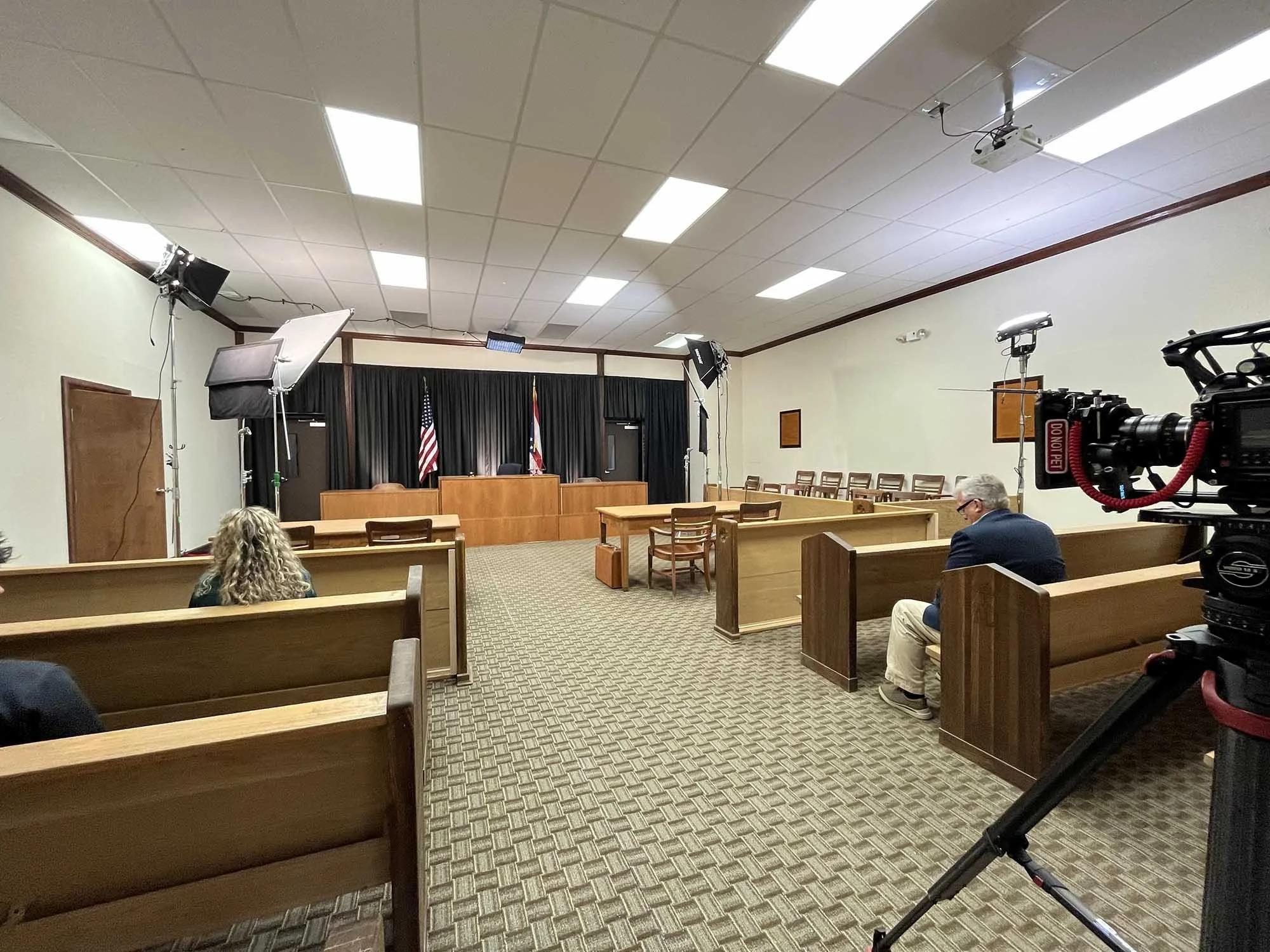 A courtroom or meeting room with wooden benches, a judge's bench, and flags at the front. There are studio lights and cameras set up, indicating filming or recording activity. Several people are seated and facing toward the front.