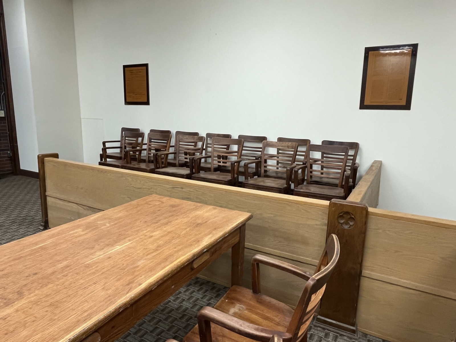 Empty waiting area with multiple wooden chairs lined up against a white wall, and a wooden table in the foreground.