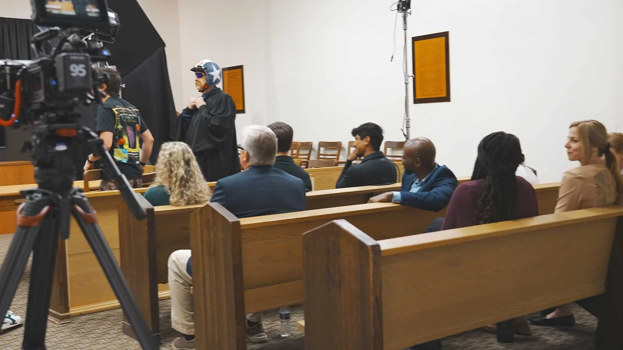People seated in wooden pews inside a courtroom, facing a person in a black gown with a helmet and sunglasses. A camera on a tripod is set up in the foreground, and a person with headphones stands near the person in gown and helmet. The room has whit