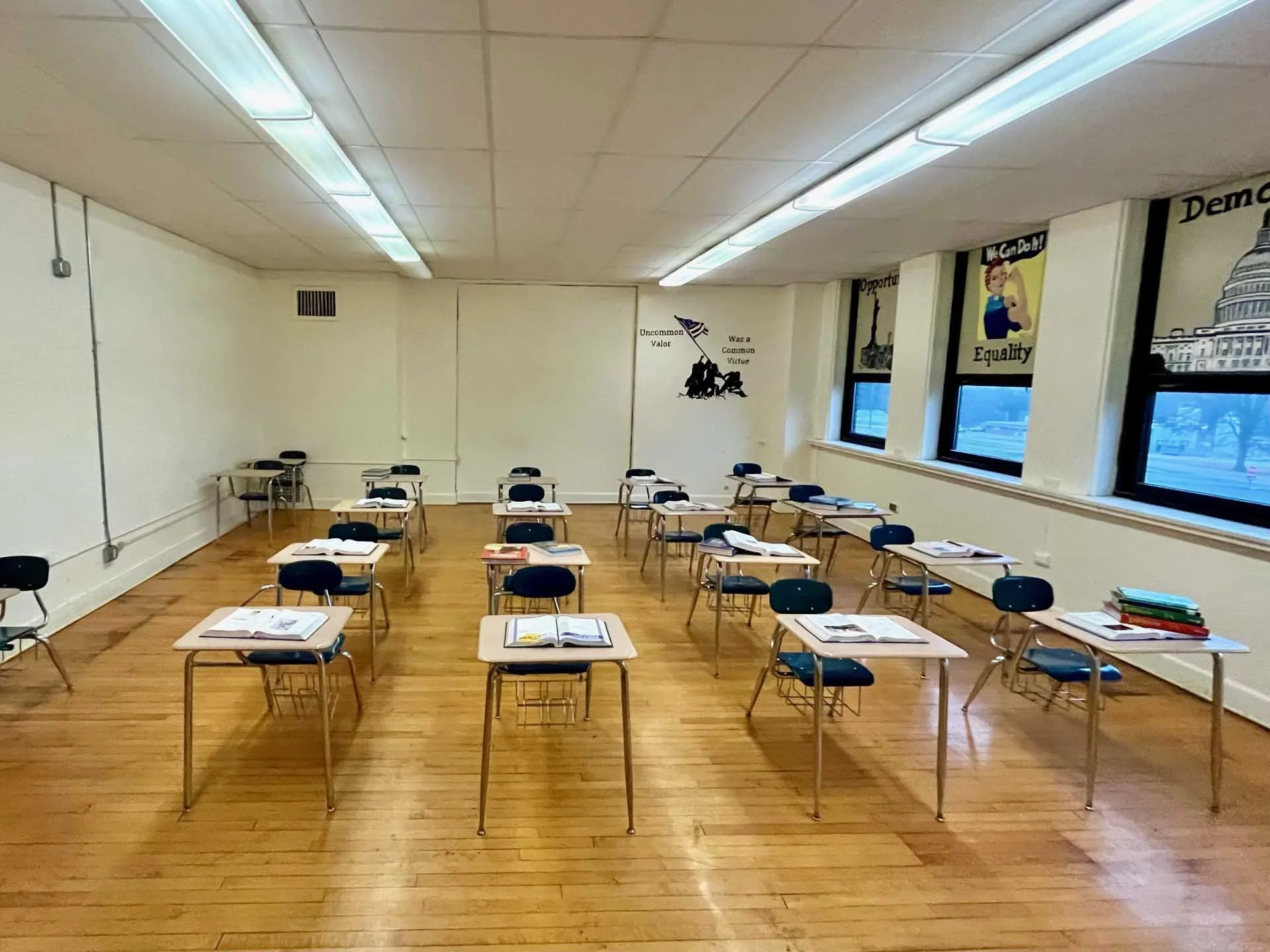 Empty classroom with several desks and chairs arranged in rows, textbooks and notebooks on the desks, windows along one wall, fluorescent lights on the ceiling, and patriotic posters on the walls.