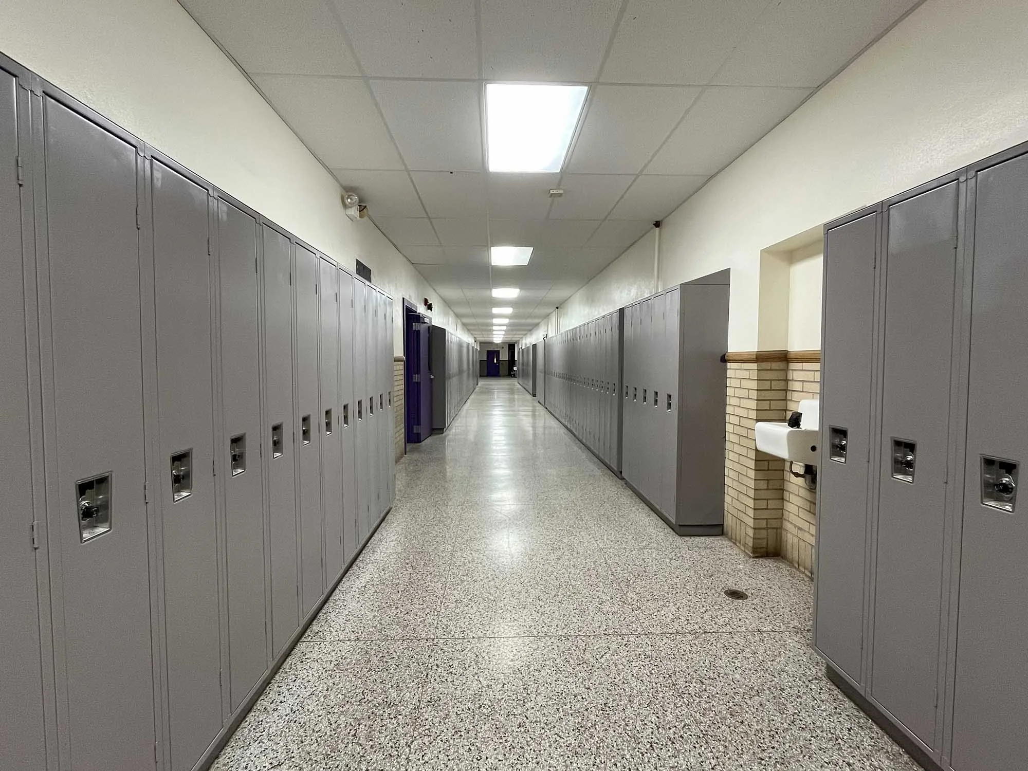 Long school hallway with gray lockers on both sides, a white sink on the right, and overhead fluorescent lighting.