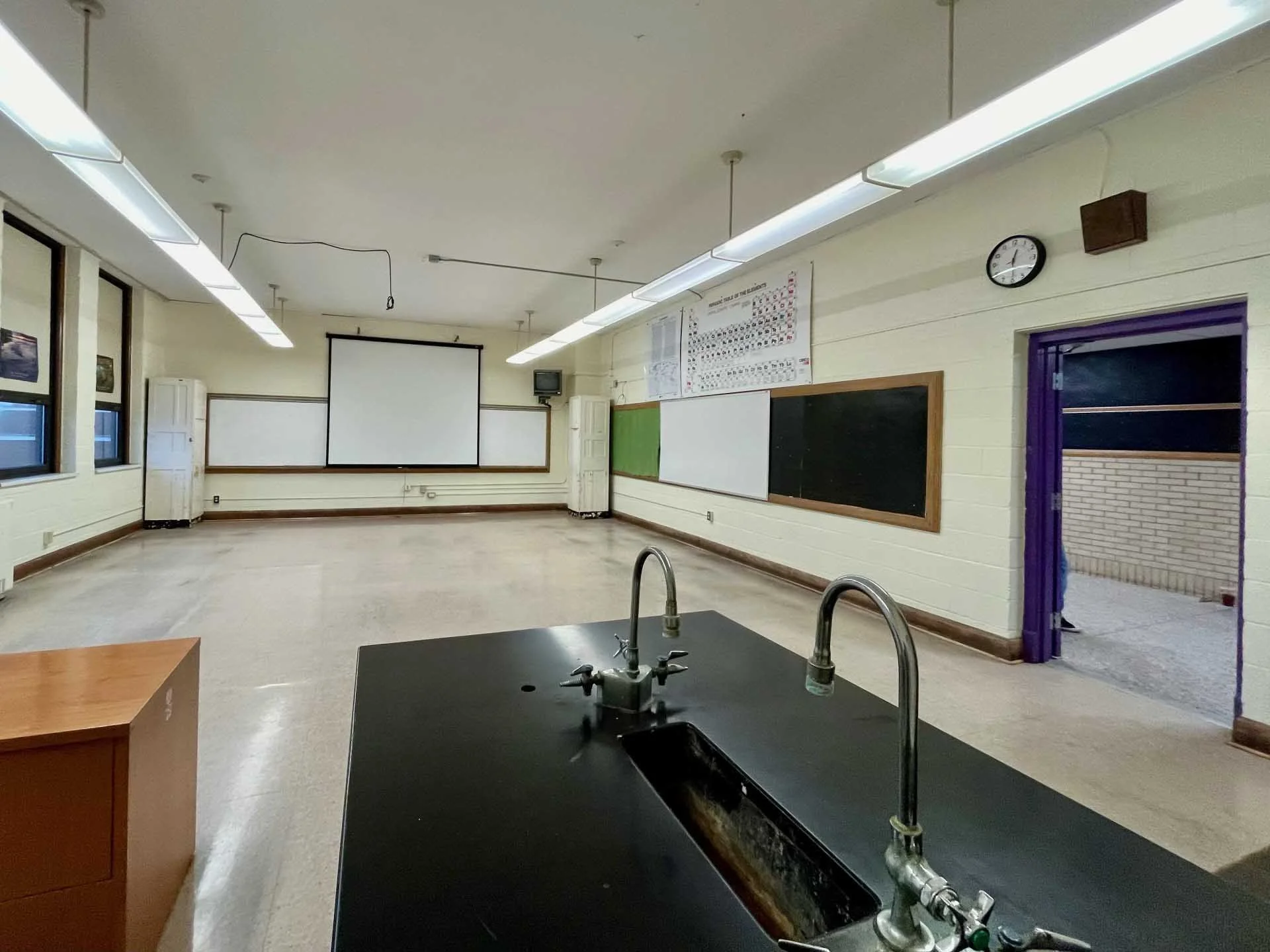 Empty science classroom with black countertop and stainless steel sinks in the foreground, whiteboard and black chalkboard on the wall, windows on the left, and a projector screen on the back wall.