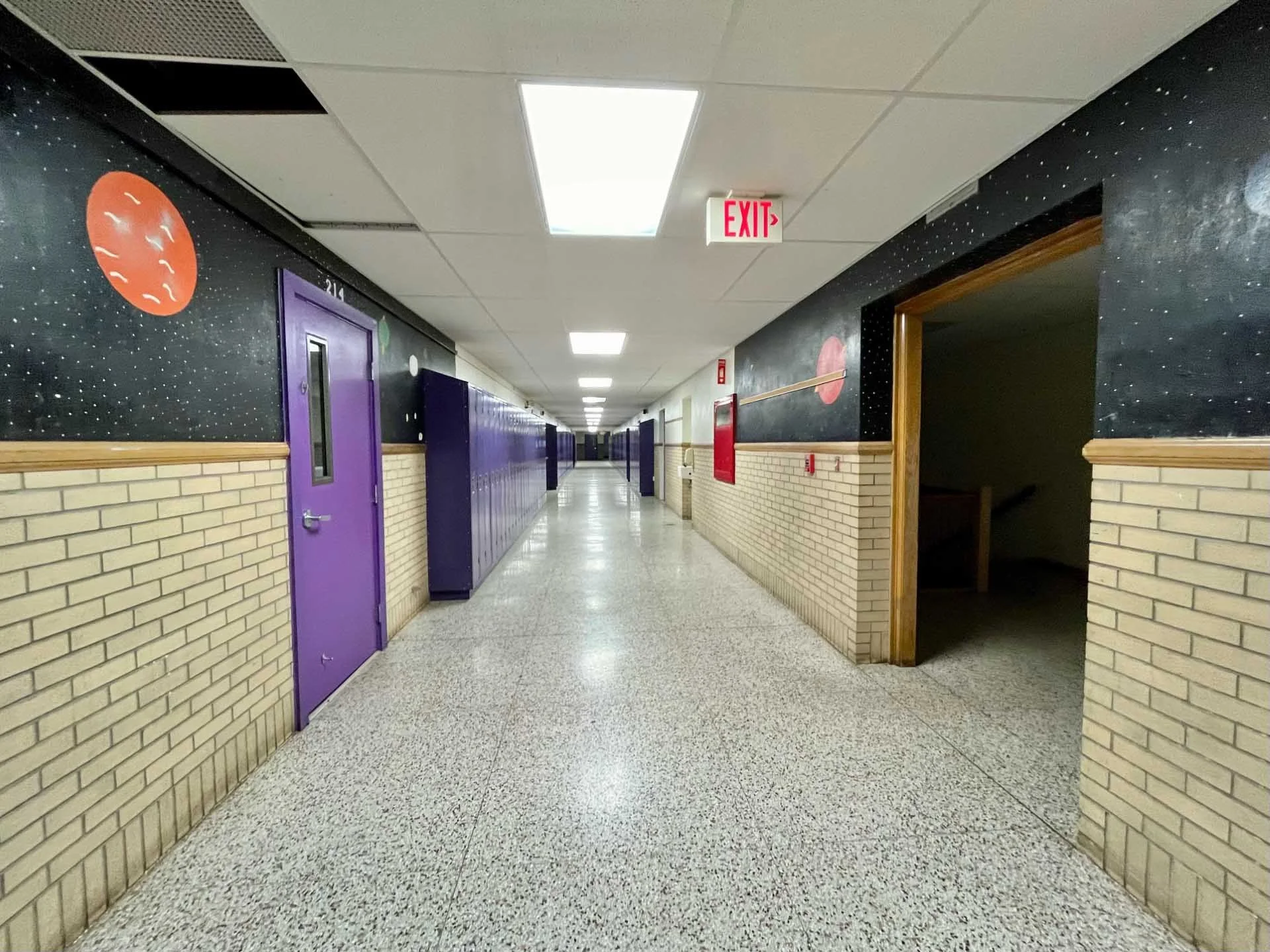 Long school hallway with purple lockers on the left and exhibit space with a door on the right, brick walls, white ceiling with fluorescent lights, and an exit sign overhead.