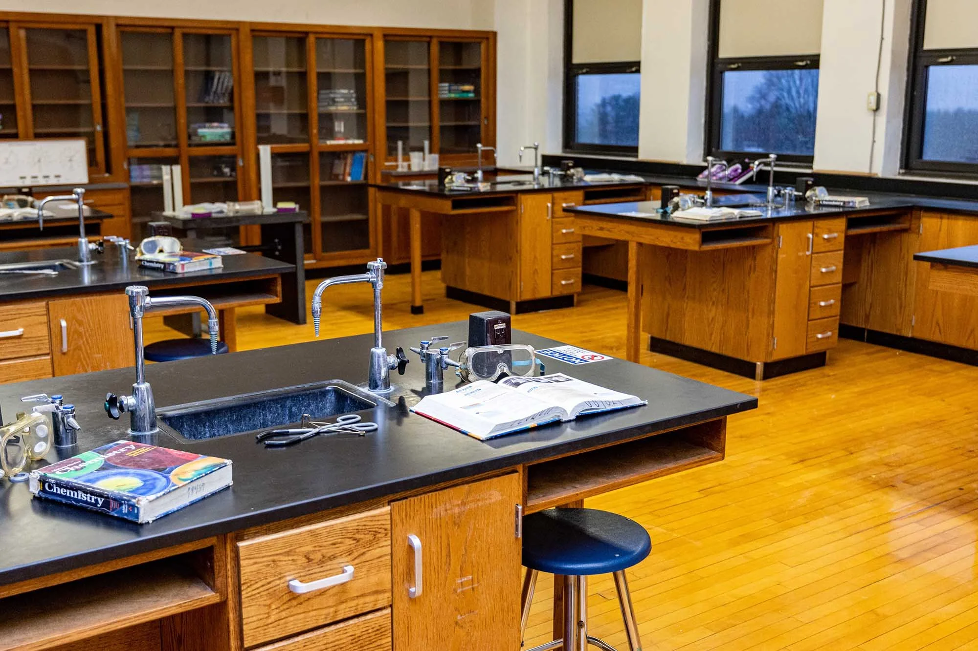 A science classroom with wooden lab benches, lab sinks, books, and large windows showing trees outside.