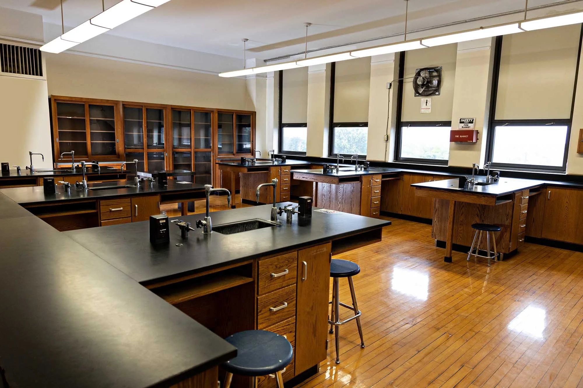 A science classroom with multiple black lab tables, sinks, and stools. Large windows along the wall let in natural light. Wooden cabinets are visible against the wall, and a wall-mounted fan and fire safety equipment are present.