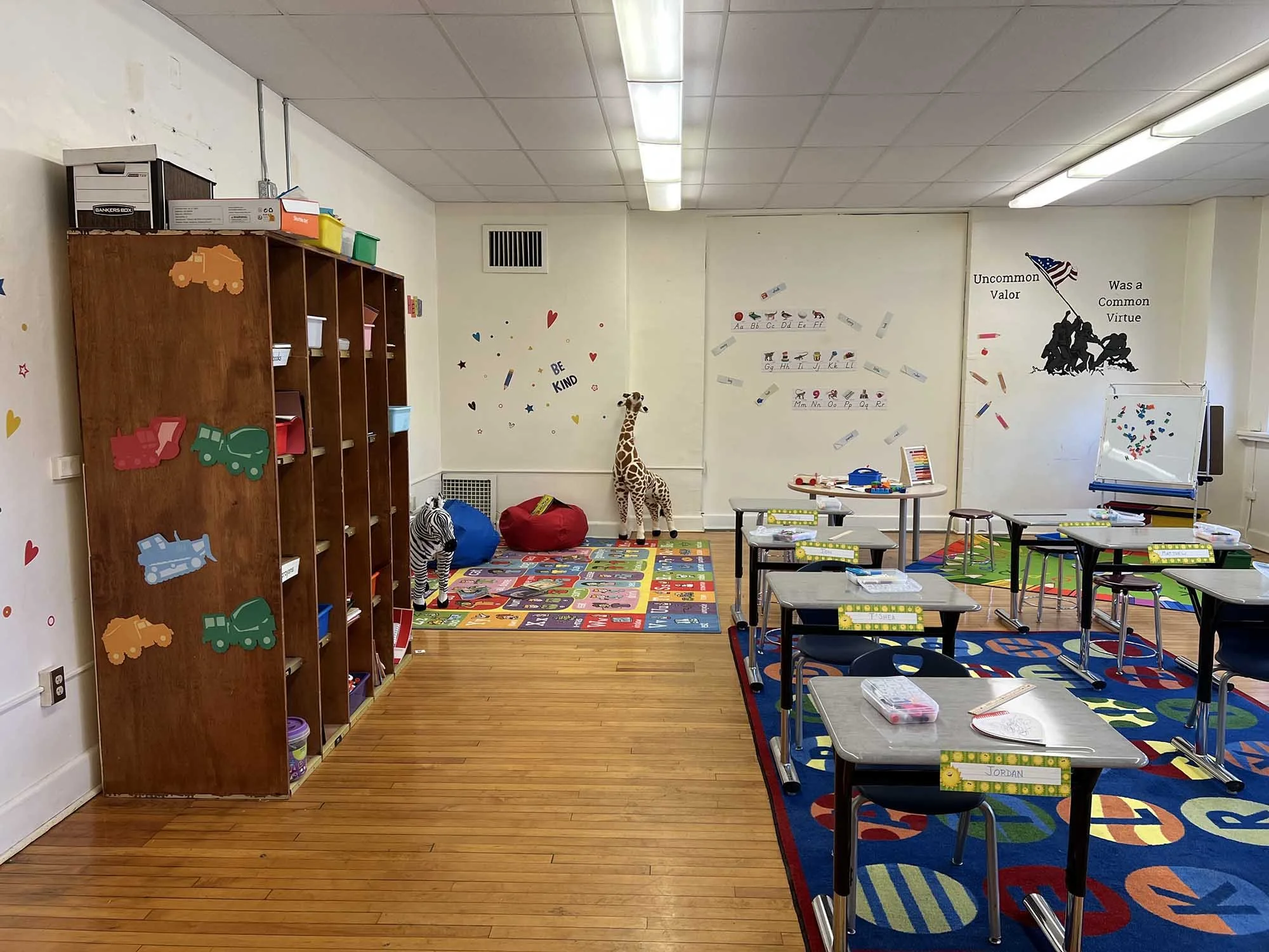 Empty preschool classroom with colorful educational decorations, a giraffe stuffed animal, small tables and chairs, and name tags on the tables.